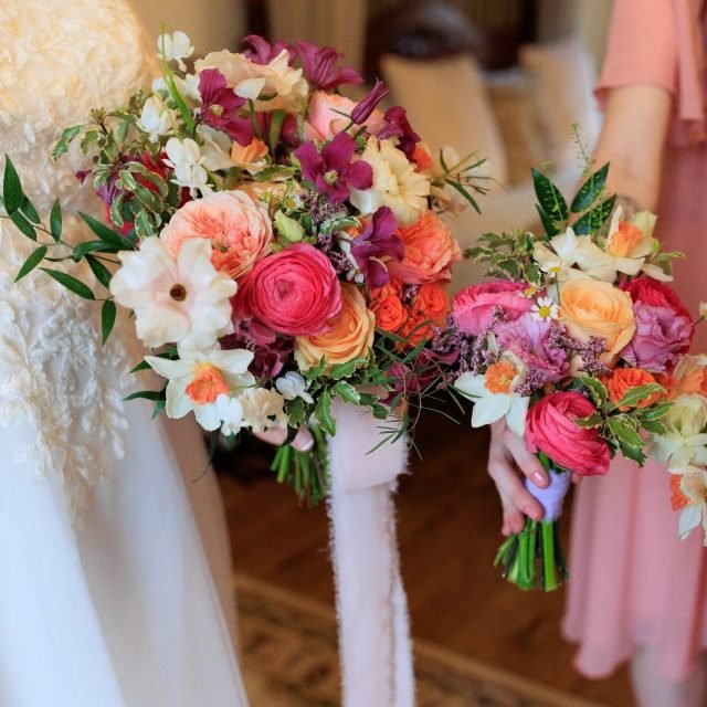 Two people holding colorful bouquets of pink, peach, white, and purple flowers at a wedding.