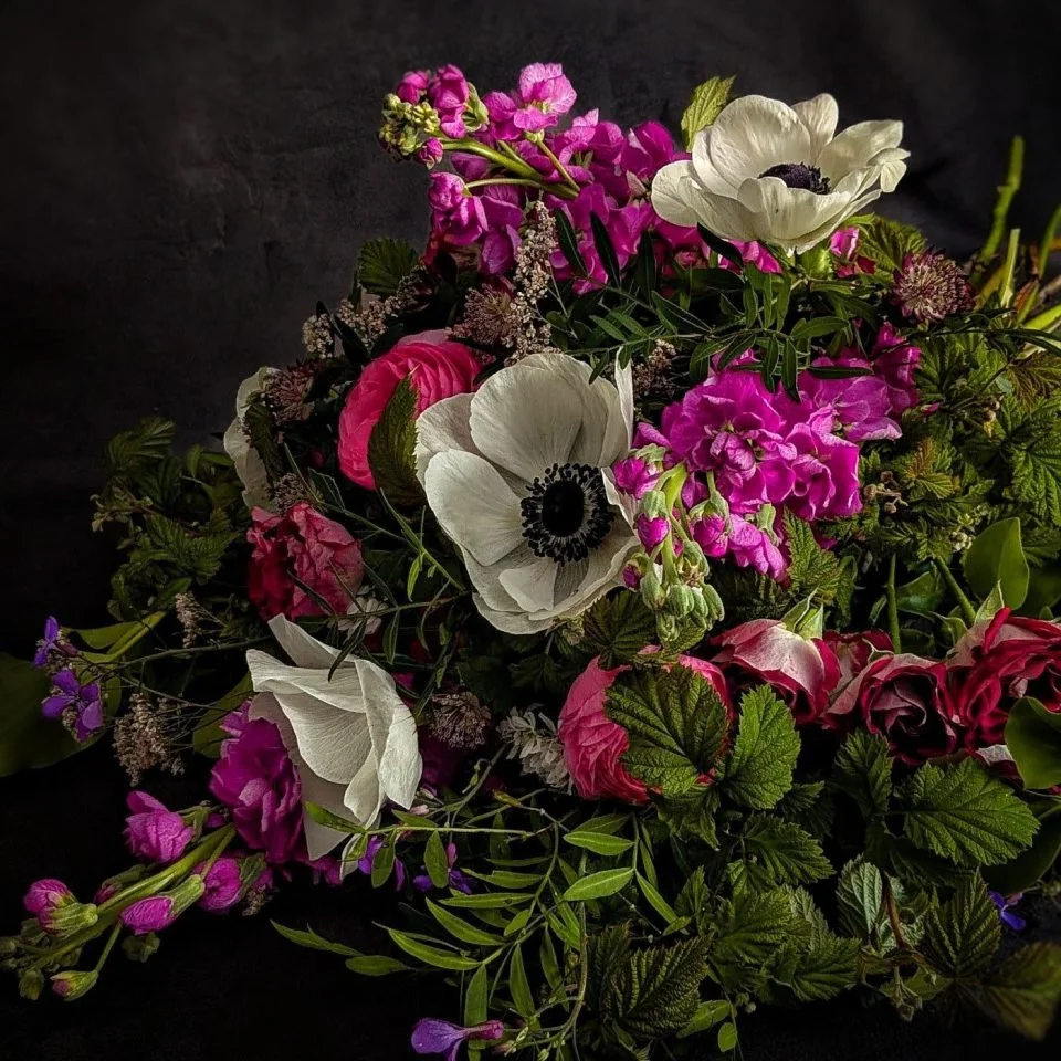 A colorful bouquet of various flowers including white anemones, pink roses, purple stock, and other small flowers and greenery against a dark background.