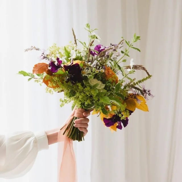 Person holding a colorful bouquet of flowers with a pink ribbon, against a white curtain background.