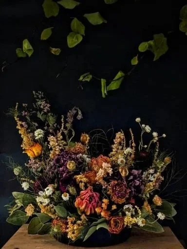 Vase of assorted colorful flowers including roses and wildflowers on a wooden surface against a dark background, with green leaves at the top.
