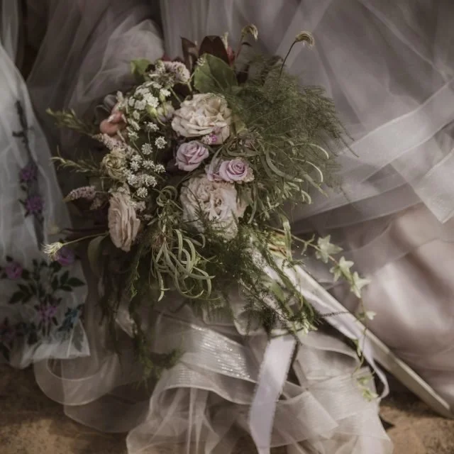 A bridal bouquet with white and pale pink roses, surrounded by greenery and white baby's breath, held by a bride in a wedding dress with tulle fabric.