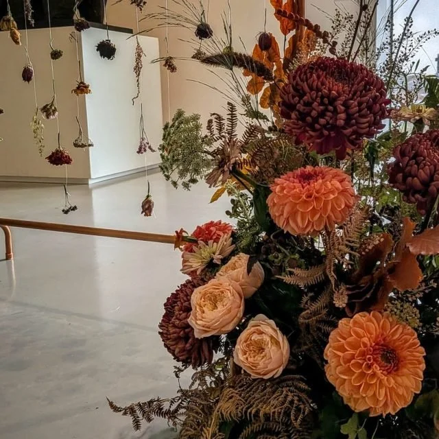 A large arrangement of orange and peach flowers, including dahlias and roses, with dried plants and foliage, in an indoor art exhibit space with hanging dried flowers in the background.