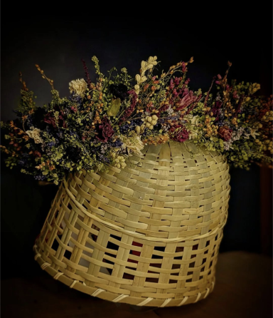 A wicker basket with a woven pattern, filled with a large arrangement of various colorful flowers and greenery, set against a dark background.