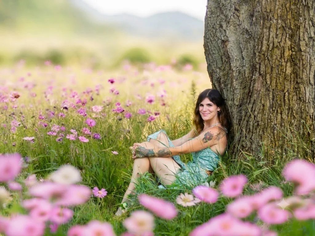 A woman with tattoos in a blue dress sitting against a tree in a field of pink and white flowers, smiling at the camera.