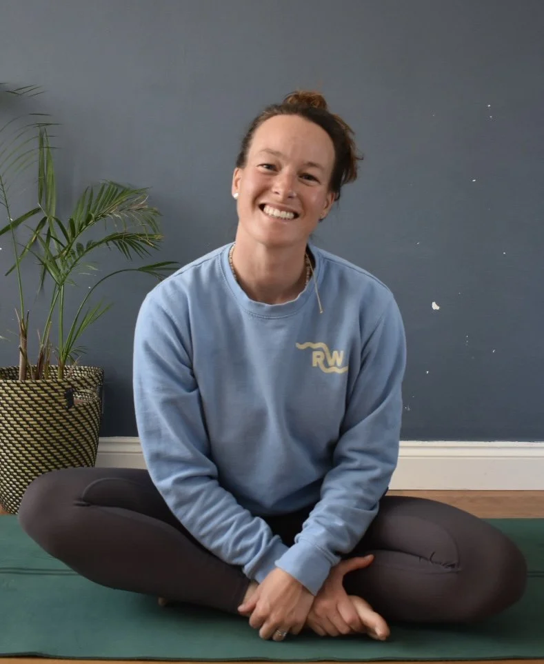A woman sitting cross-legged on a yoga mat with a smile, wearing a light blue sweatshirt, inside a room with a dark blue wall and a potted plant nearby.