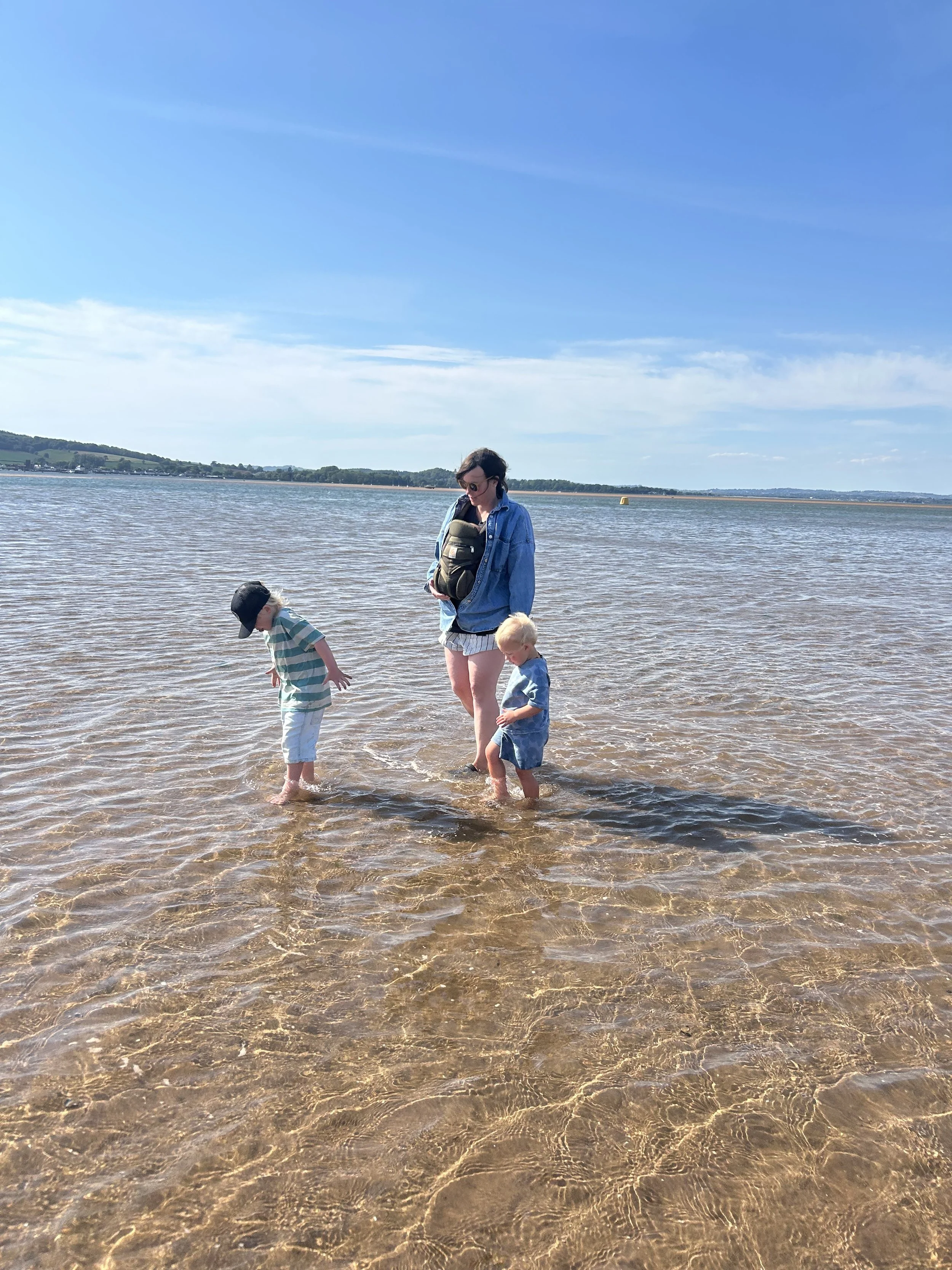 A woman and three children standing in shallow water by a lake on a sunny day, with a blue sky and distant shoreline in the background.