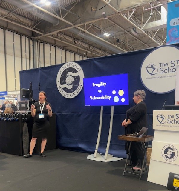 Two women giving a presentation at an indoor event or conference. One woman is speaking in front of a screen that displays a slide titled 'Fragility vs Vulnerability'. The background shows a large banner with a logo for The ST School and Hands on Hub, indicating an educational or professional development setting.