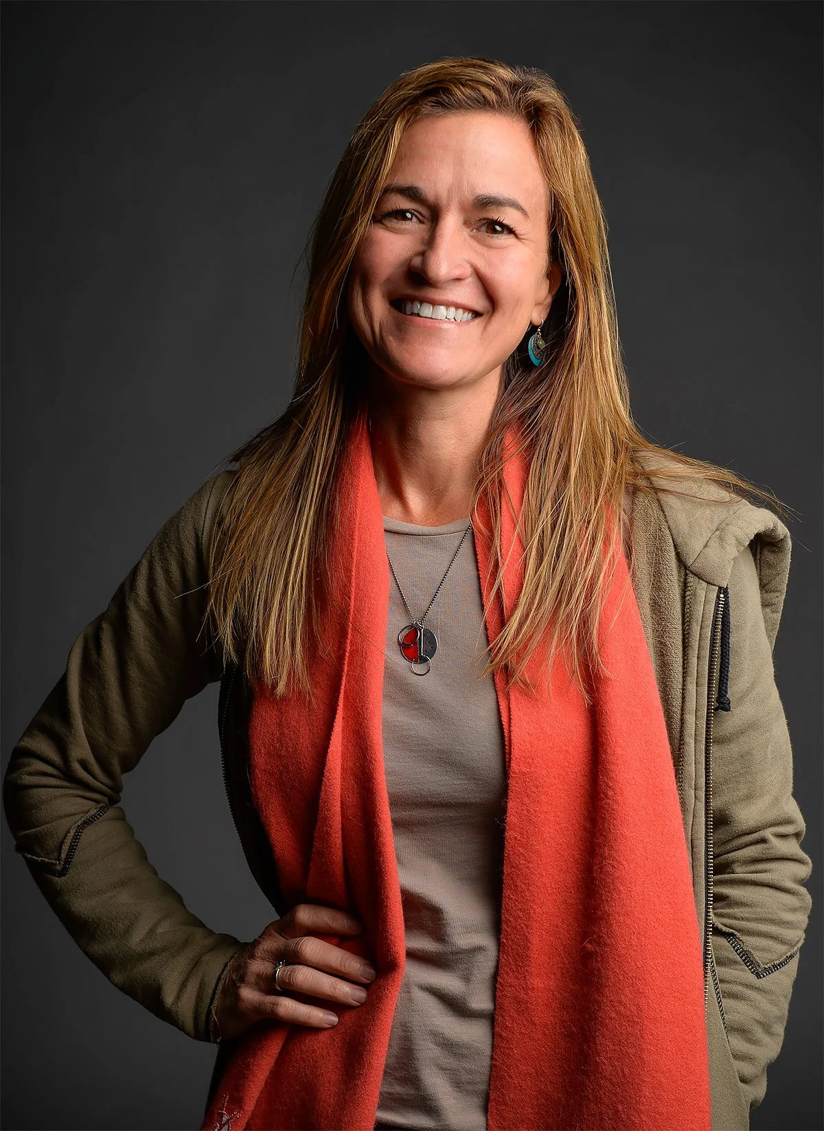 A woman with long, straight, light brown hair, smiling, wearing a beige shirt, a tan jacket, a red scarf, and jewelry including a yin-yang necklace and turquoise earrings, standing against a dark gray background.