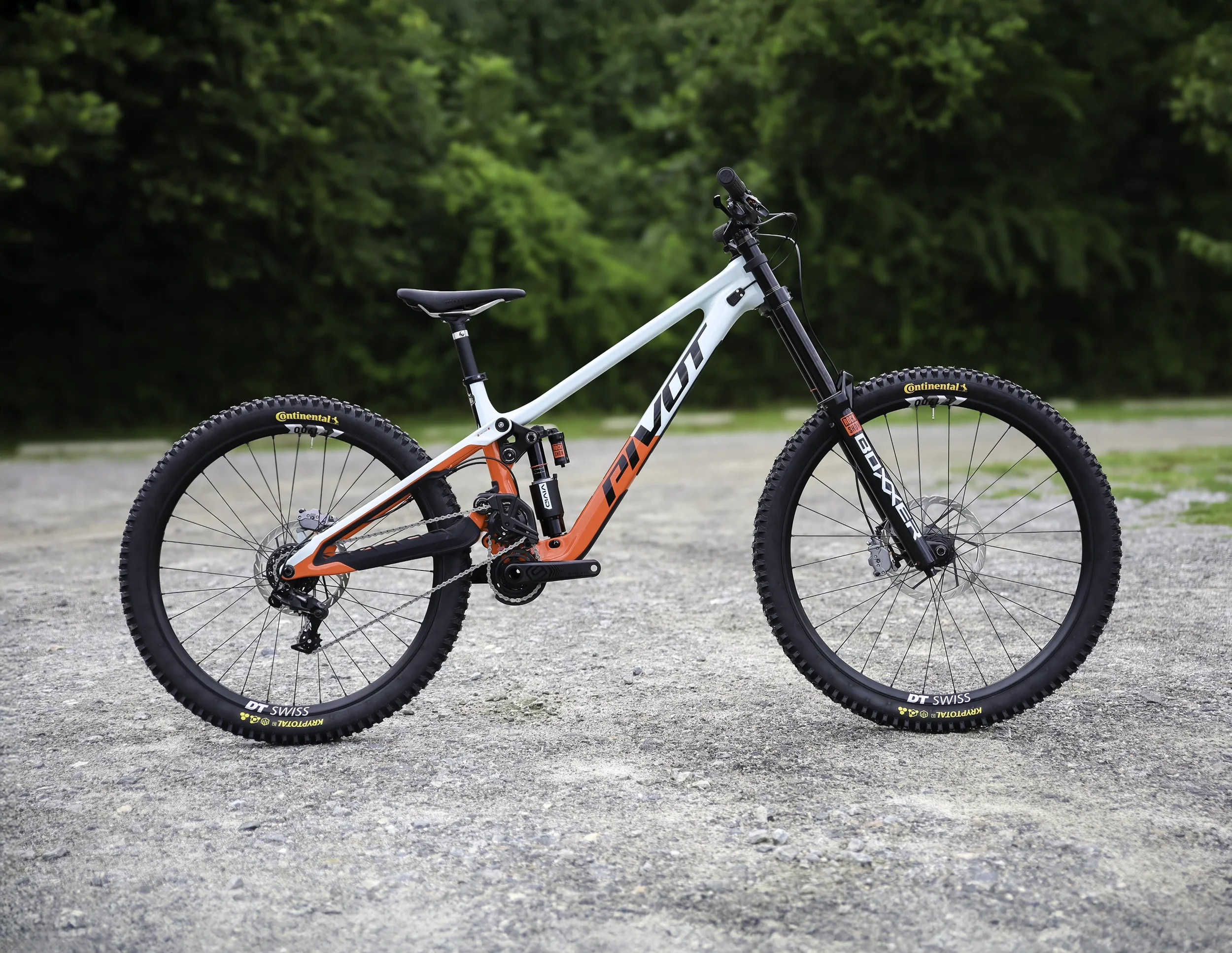 A modern mountain bike with large knobby tires and a light blue and red frame, parked on a gravel surface in front of a background of green trees.
