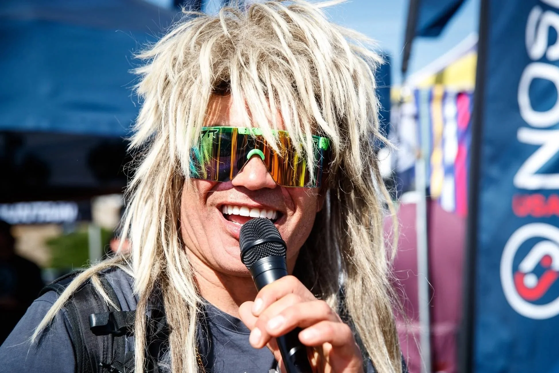 Brett Tippie long, messy blonde hair wearing reflective sunglasses and a dark jacket, speaking into a microphone at Sea Otter Classic 2023, smiling. There are tents and banners in the background.
