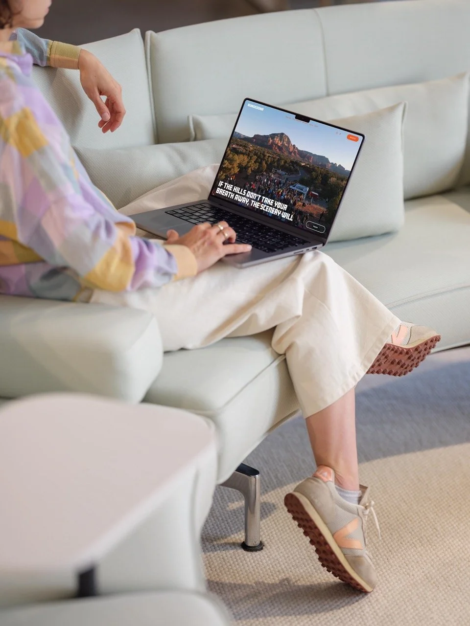Person sitting on a cream-colored sofa using a laptop, wearing beige sneakers and cream pants, with a multi-colored top partially visible.