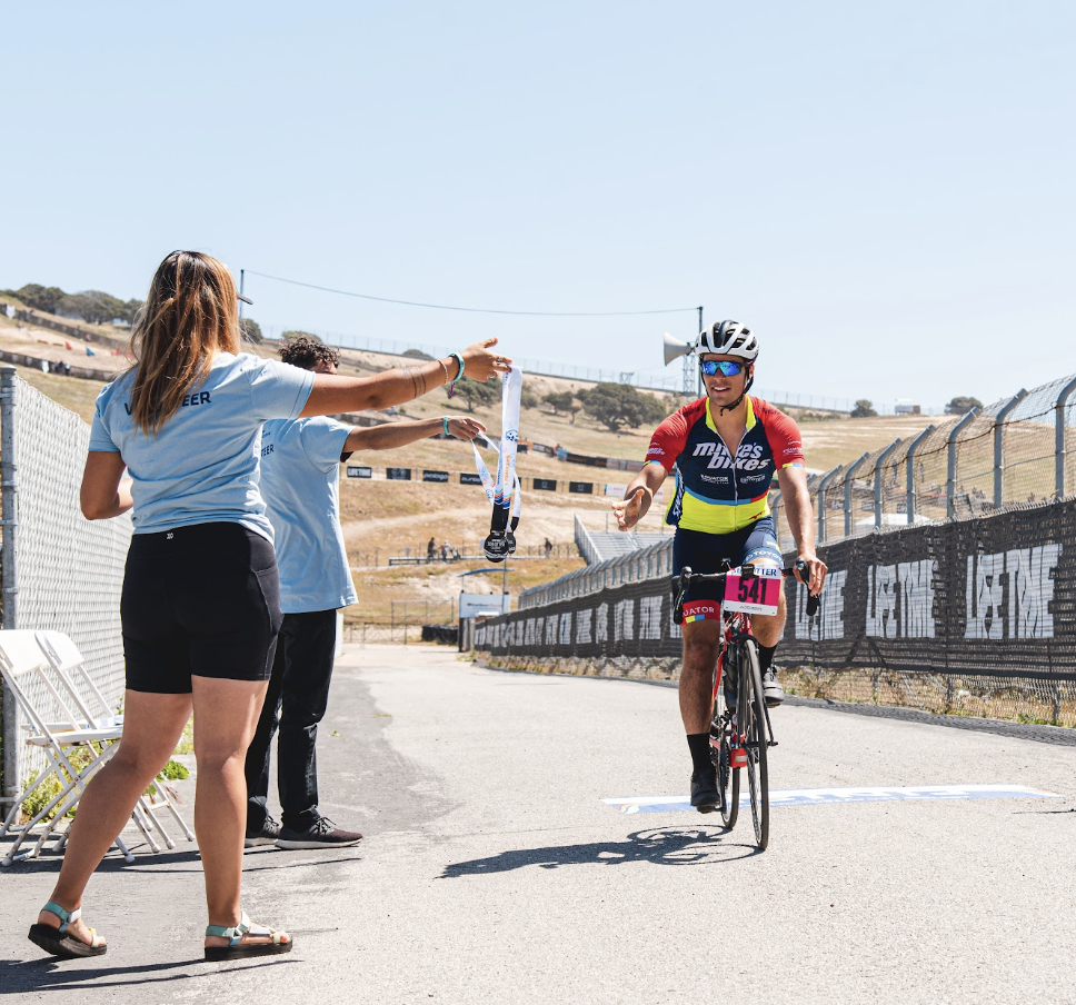 A cyclist crossing the finish line of a race on a sunny day, receiving a medal from volunteers.