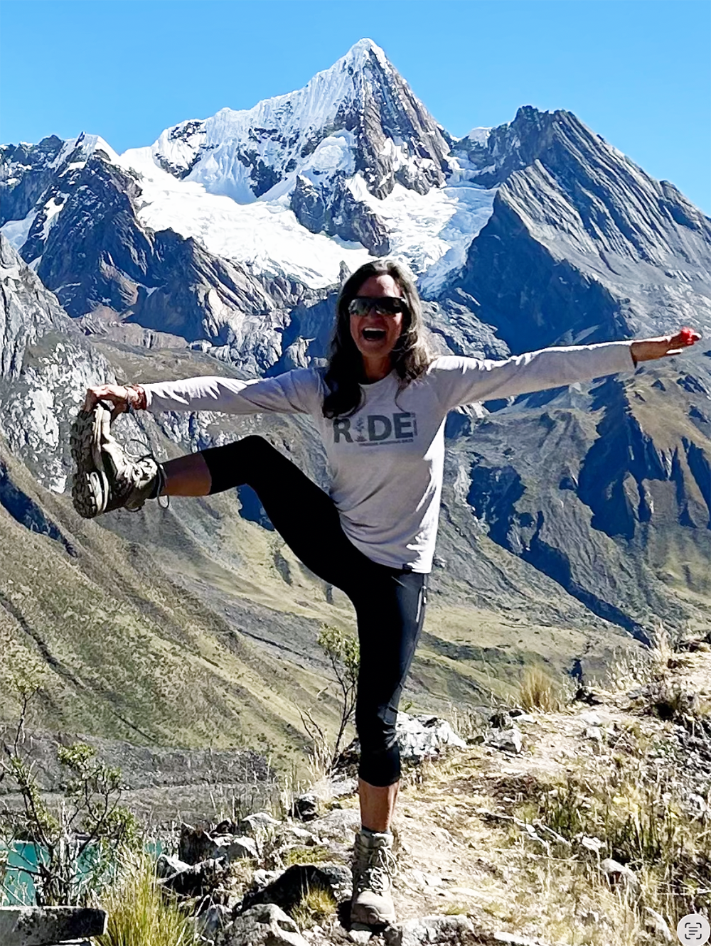 A woman doing a yoga pose with arms extended and one leg raised on a mountain trail, with snow-capped mountains and blue sky in the background.
