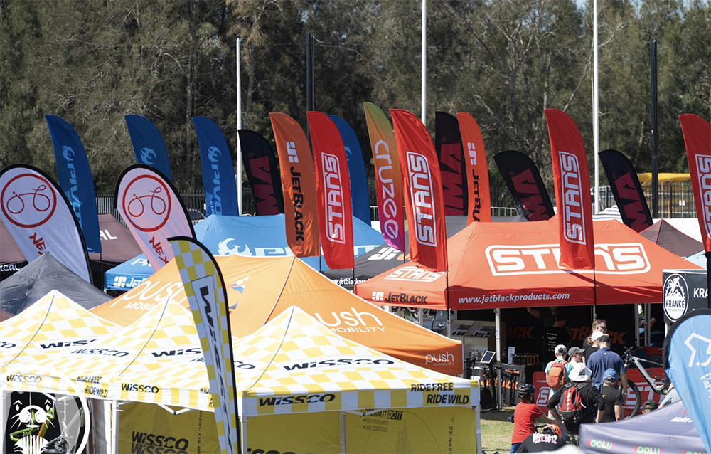 Outdoor scene with numerous tents and flags at a cycling event, featuring logos of brands like WISSO, USH, STANS, and JetBlack, with people walking and interacting.