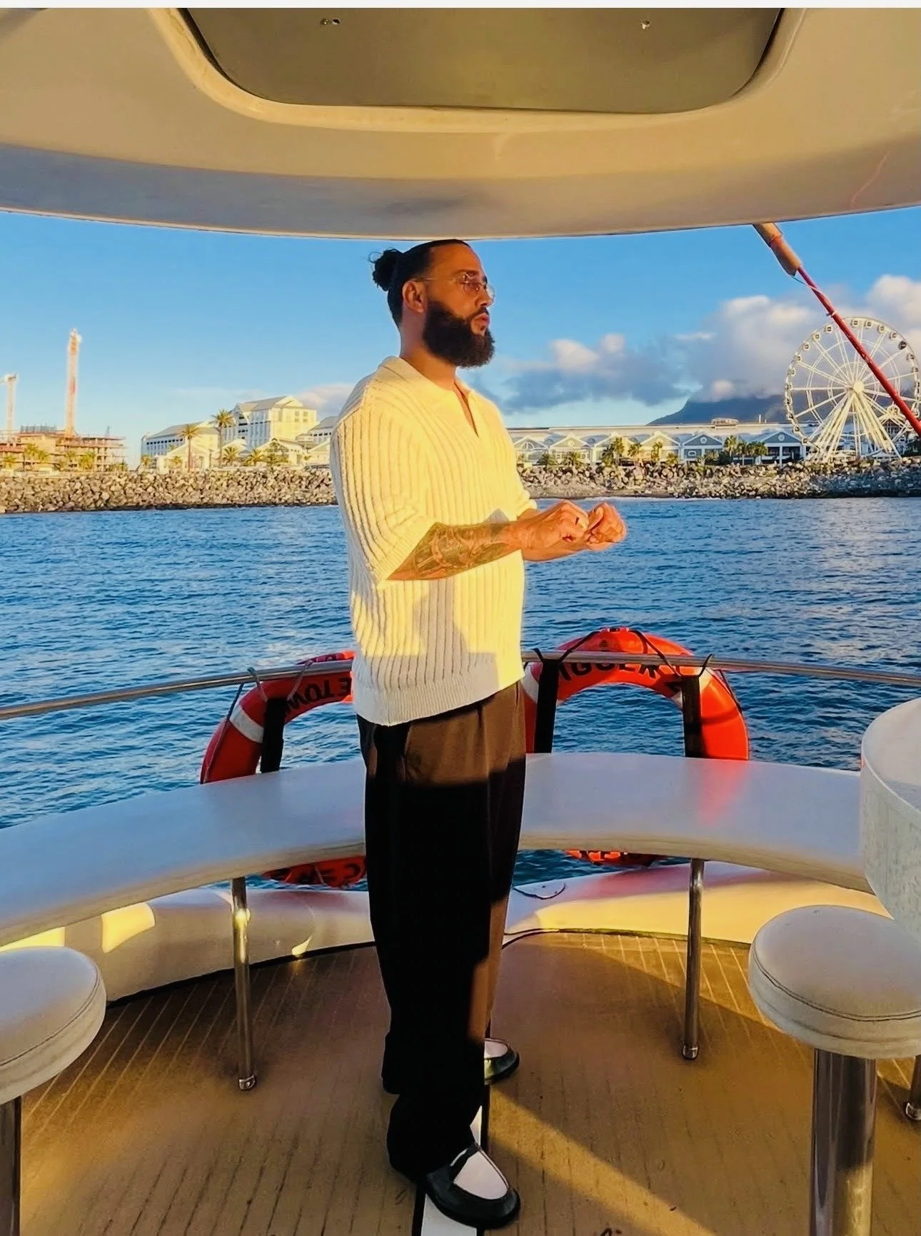 A man with a beard, glasses, and a bun hairstyle standing on a boat, facing to the right, with water and a cityscape with a Ferris wheel in the background.
