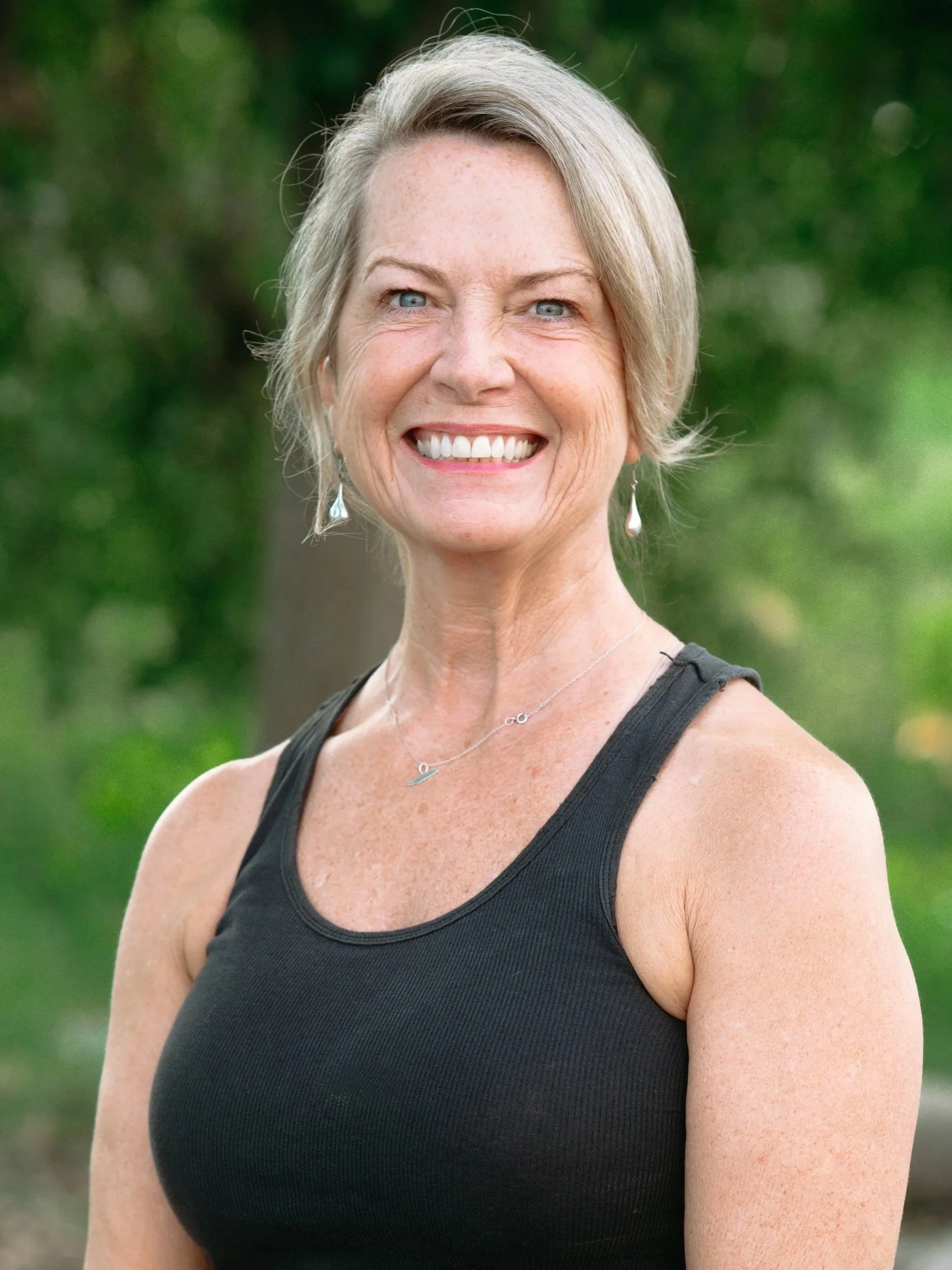 A smiling woman with short gray hair, wearing a black sleeveless top, earrings, and a necklace, outdoors with green blurred background.