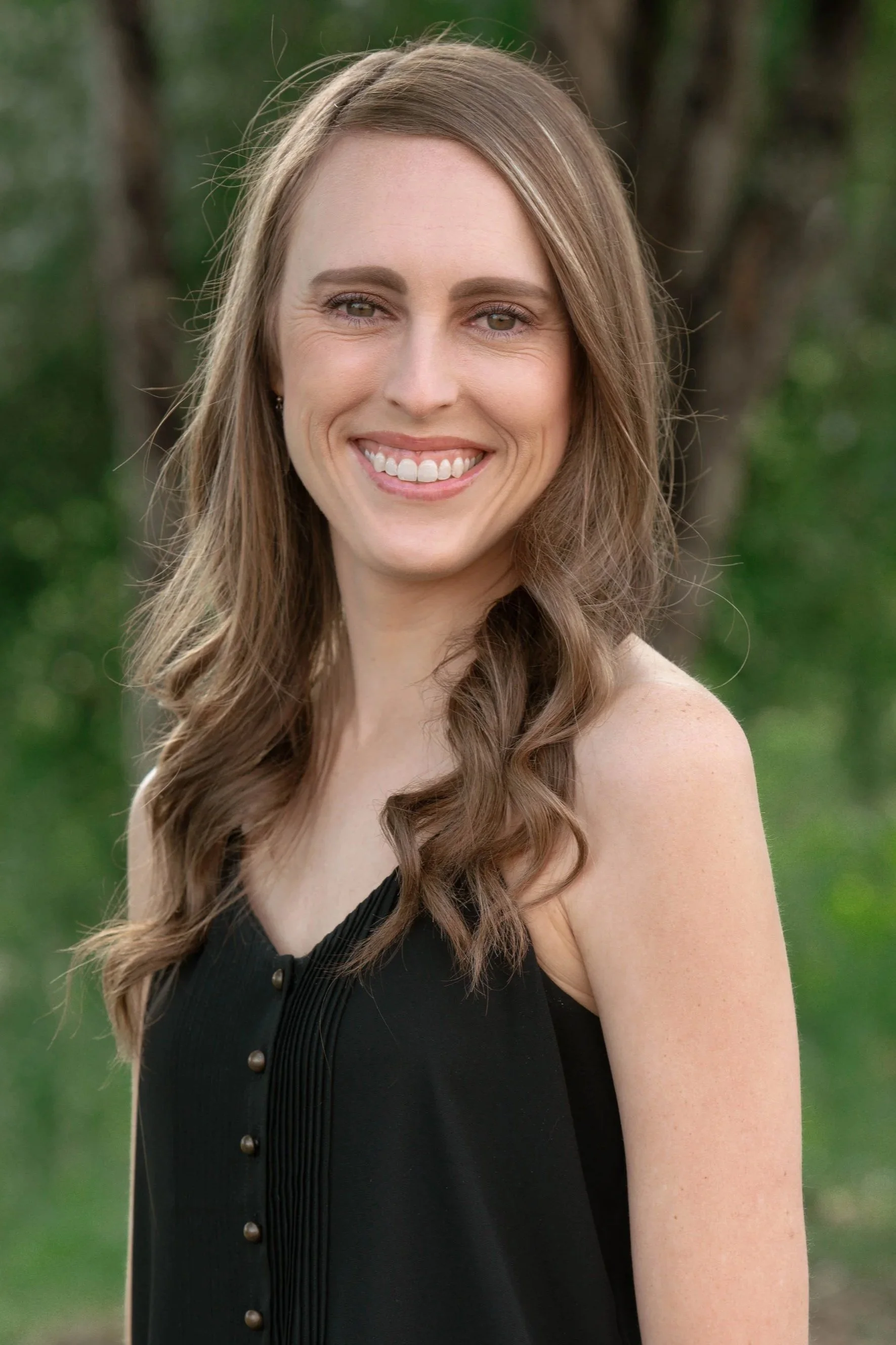A woman with long wavy brown hair, wearing a black dress, smiling outdoors with green blurred trees in the background.