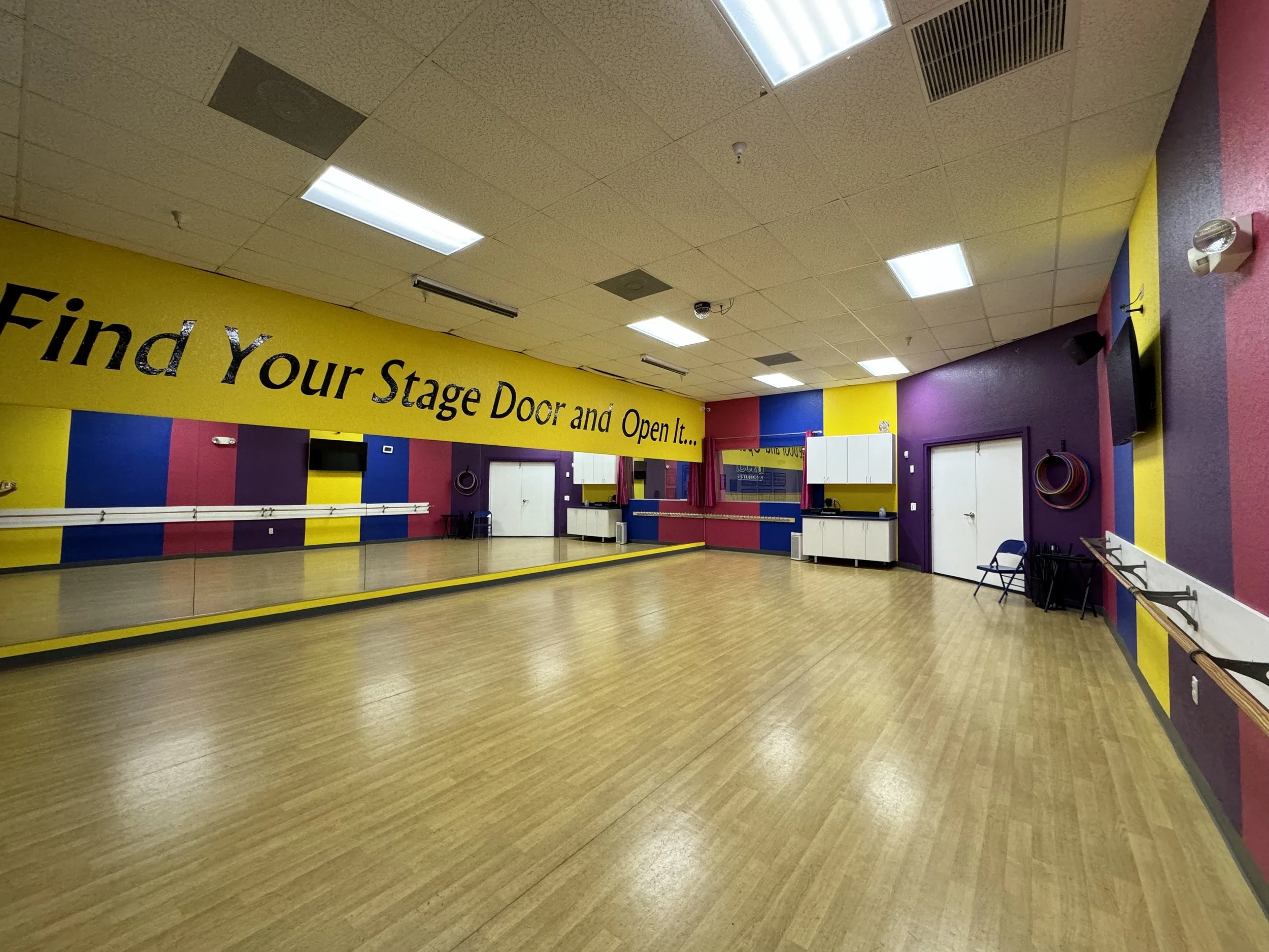 A colorful dance studio with a wood floor, purple, yellow, pink, and blue walls, a mirror, and a yellow banner that says "Find Your Stage Door and Open It...".
