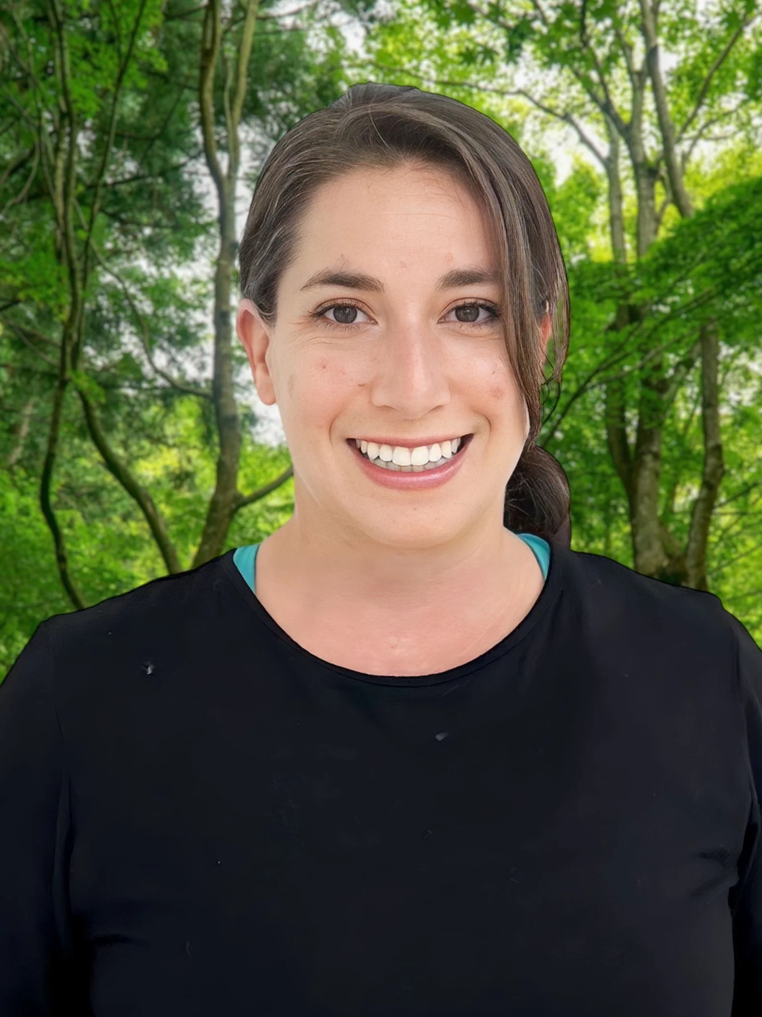 A smiling woman with brown hair and blue eyes, wearing a black shirt, standing outdoors in a green forested area with trees and foliage in the background.
