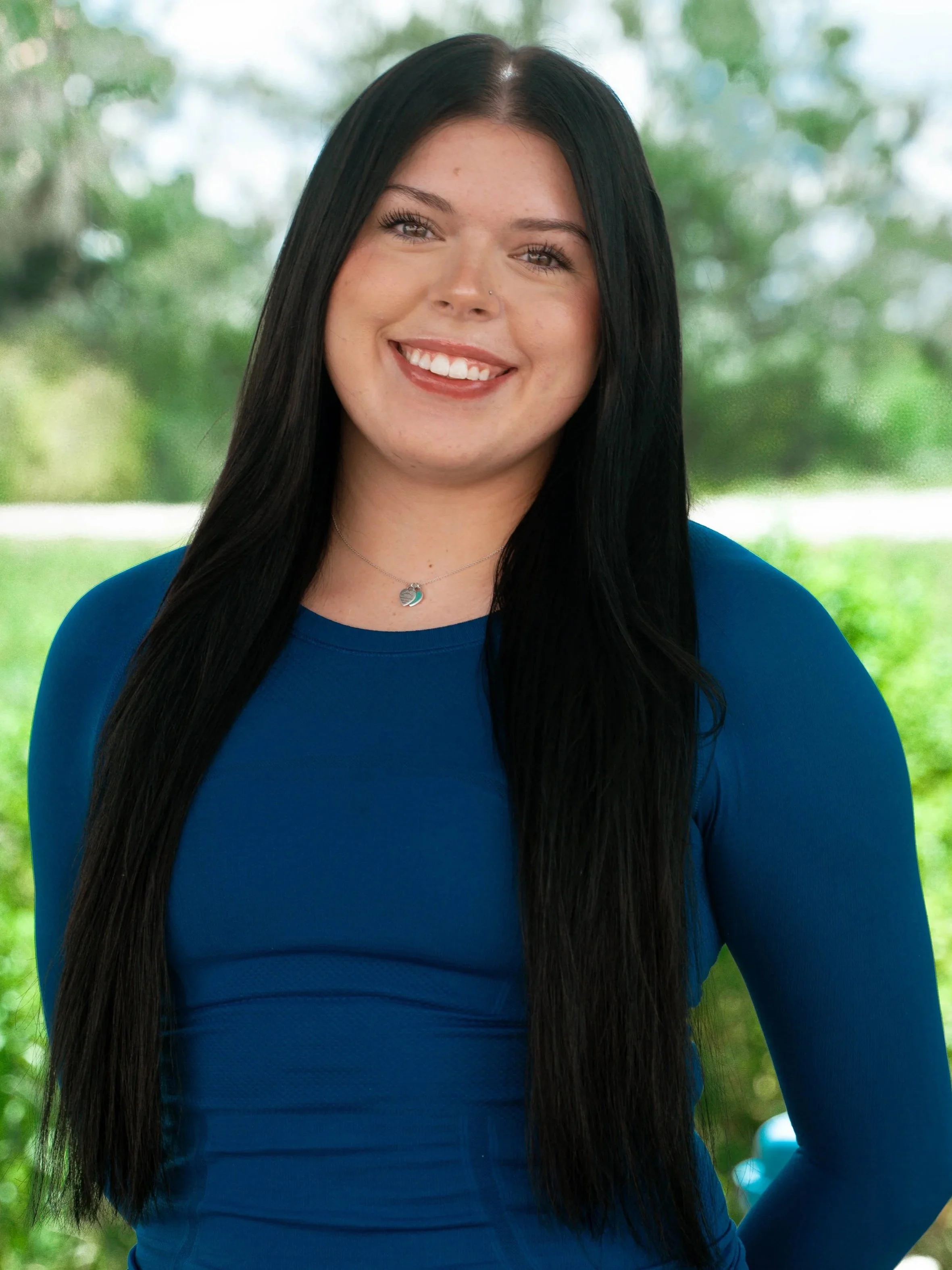 A young woman with long black hair, smiling, wearing a blue top and a necklace, outdoors with a blurred green background.