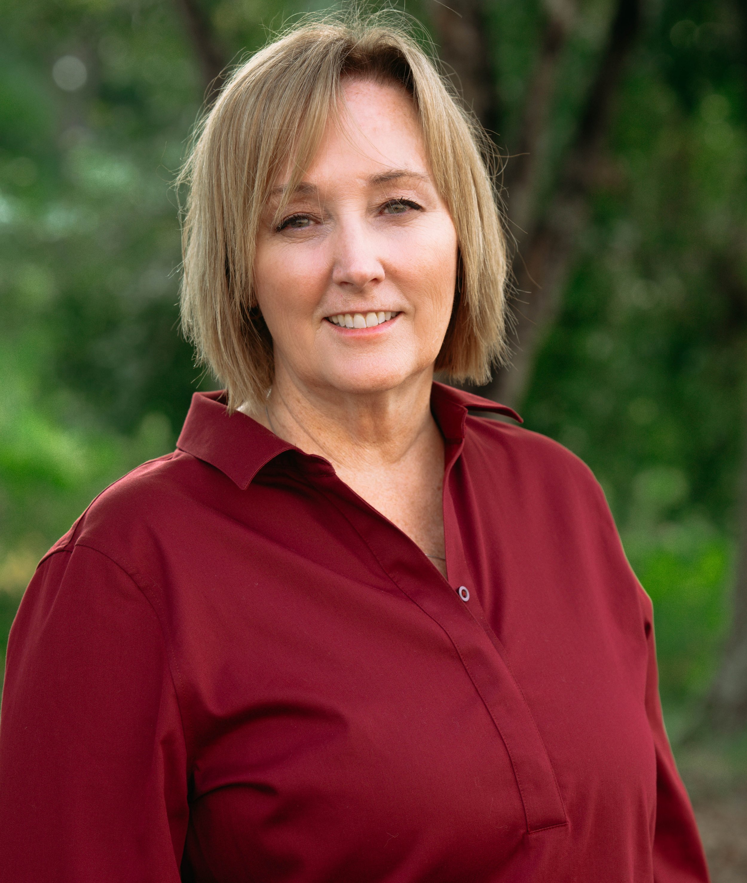 Close-up of a middle-aged woman with blonde hair wearing a burgundy shirt outdoors with a blurred green background.
