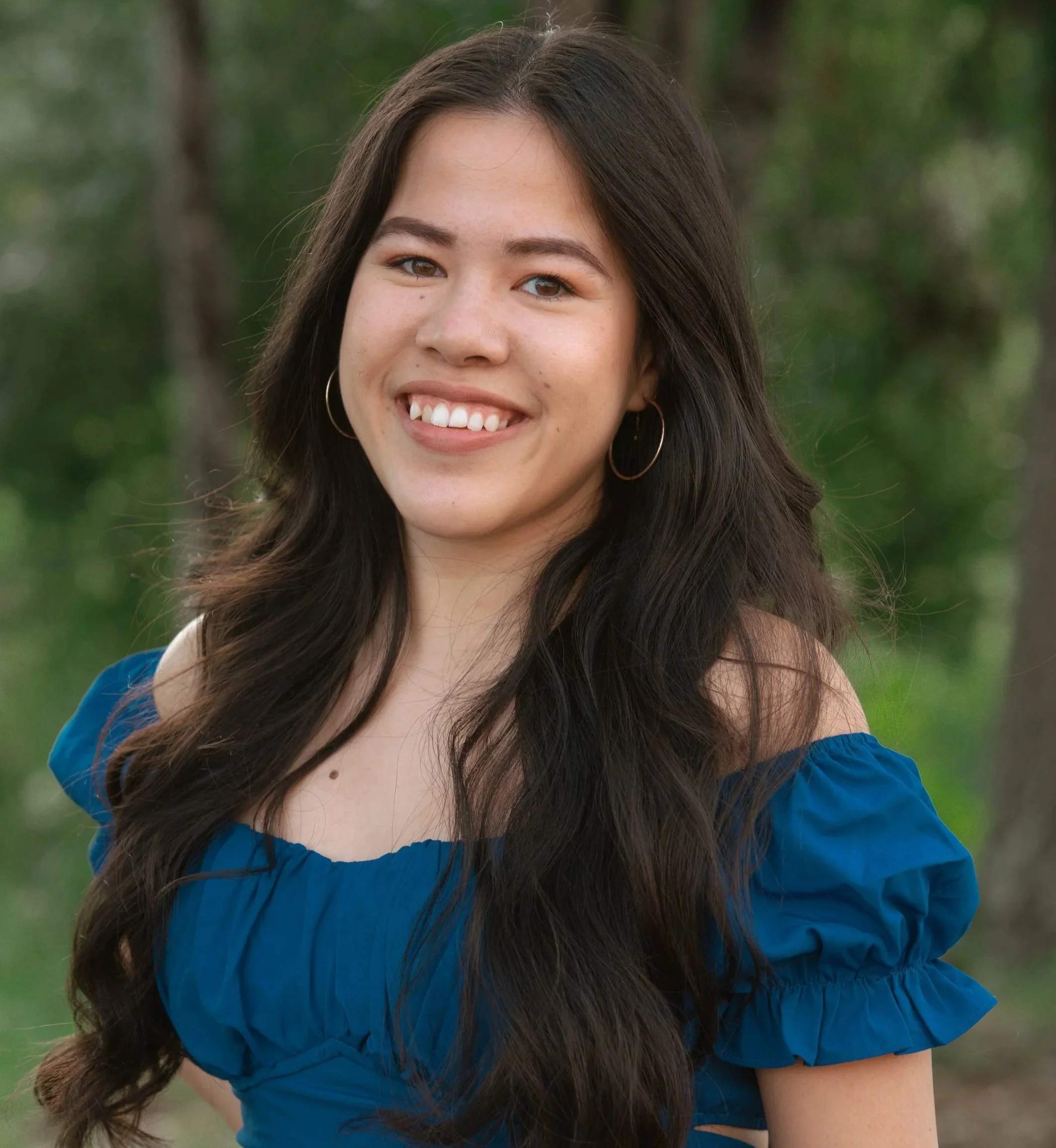 Smiling young woman with long dark hair wearing a blue off-the-shoulder top standing outdoors with blurred trees in the background.