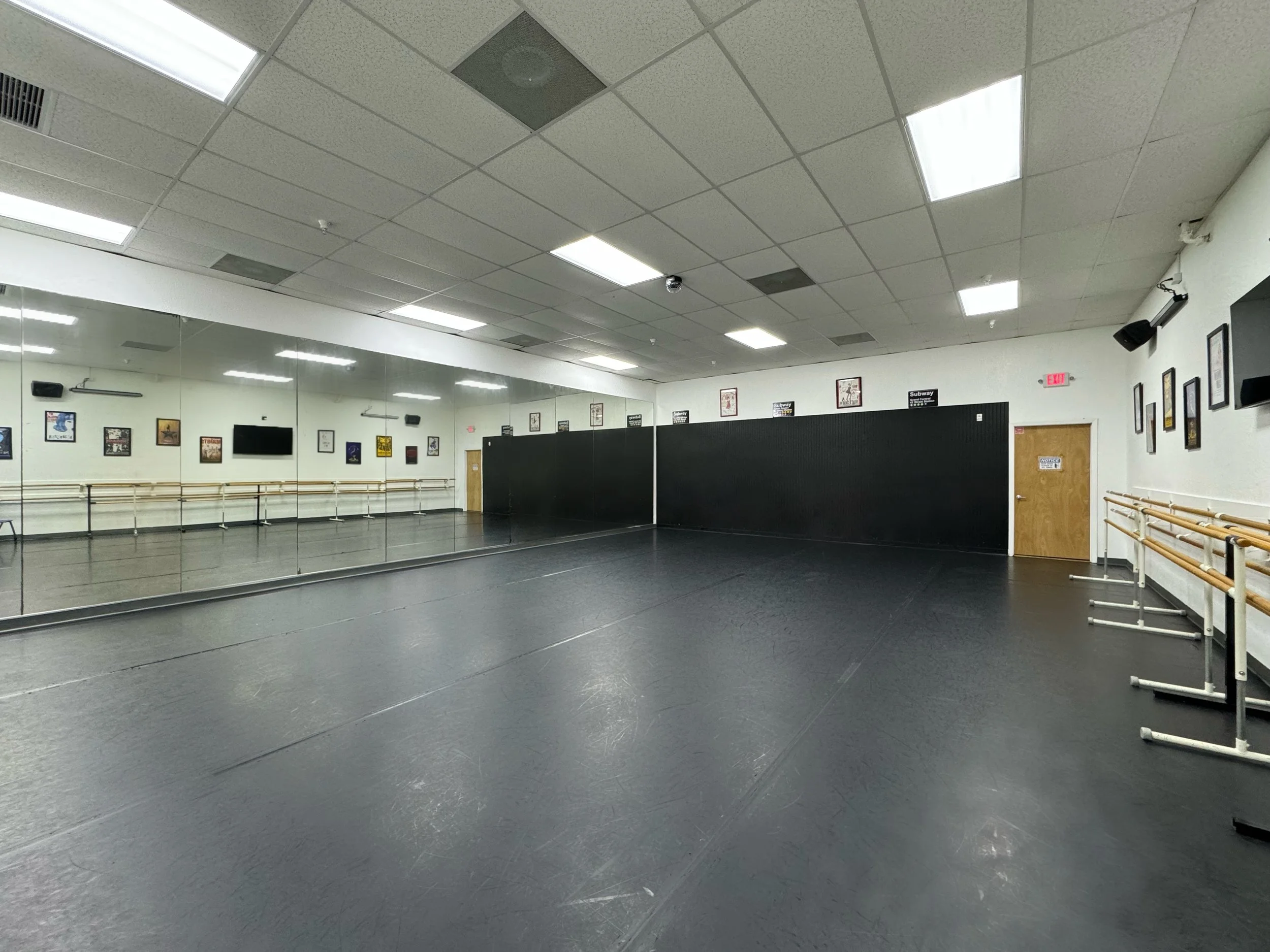 Empty dance studio with large mirrors on wall, ballet barres, black floor, and framed pictures on the wall.