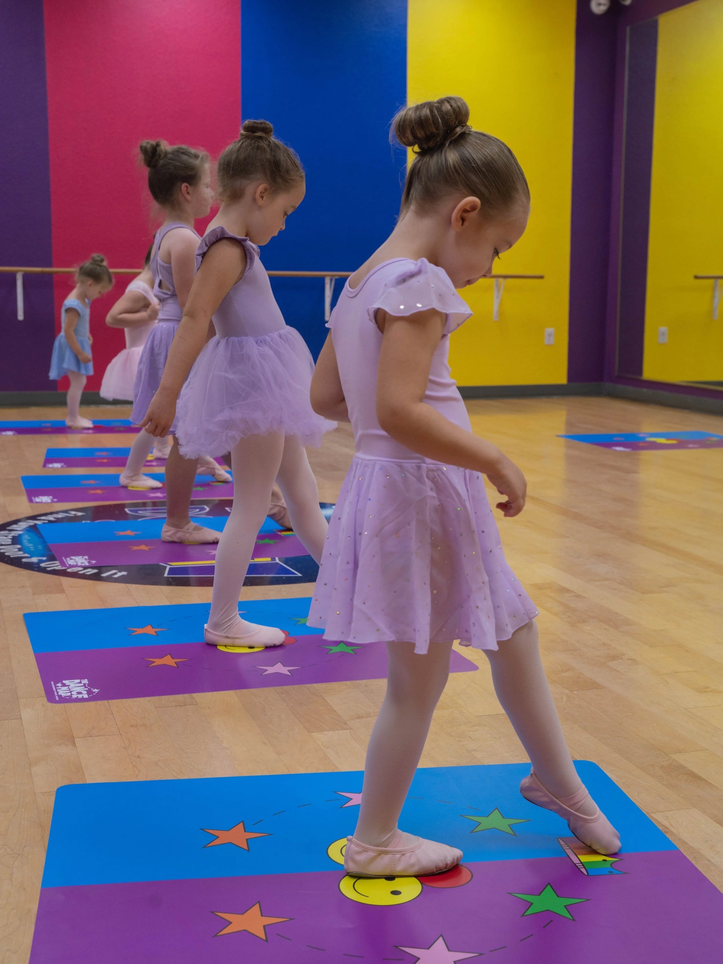 Four young girls in ballet tutus practicing ballet in a dance studio with colorful walls, standing on colorful mats.