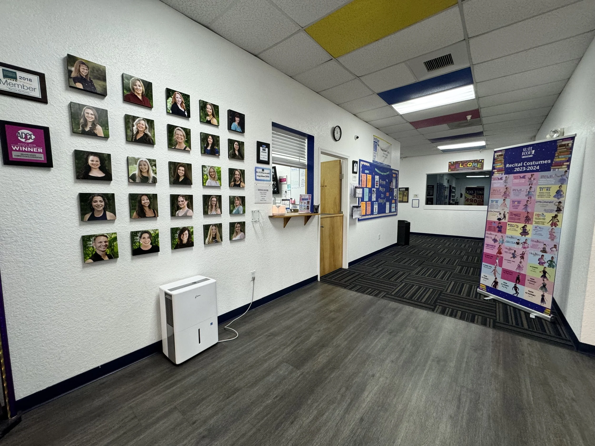 The image shows a hallway with a wall decorated with numerous framed photographs of women, likely members or students, arranged in a grid. There are various informational and decorative posters, a clock on the wall, and a stand with pamphlets. The ce