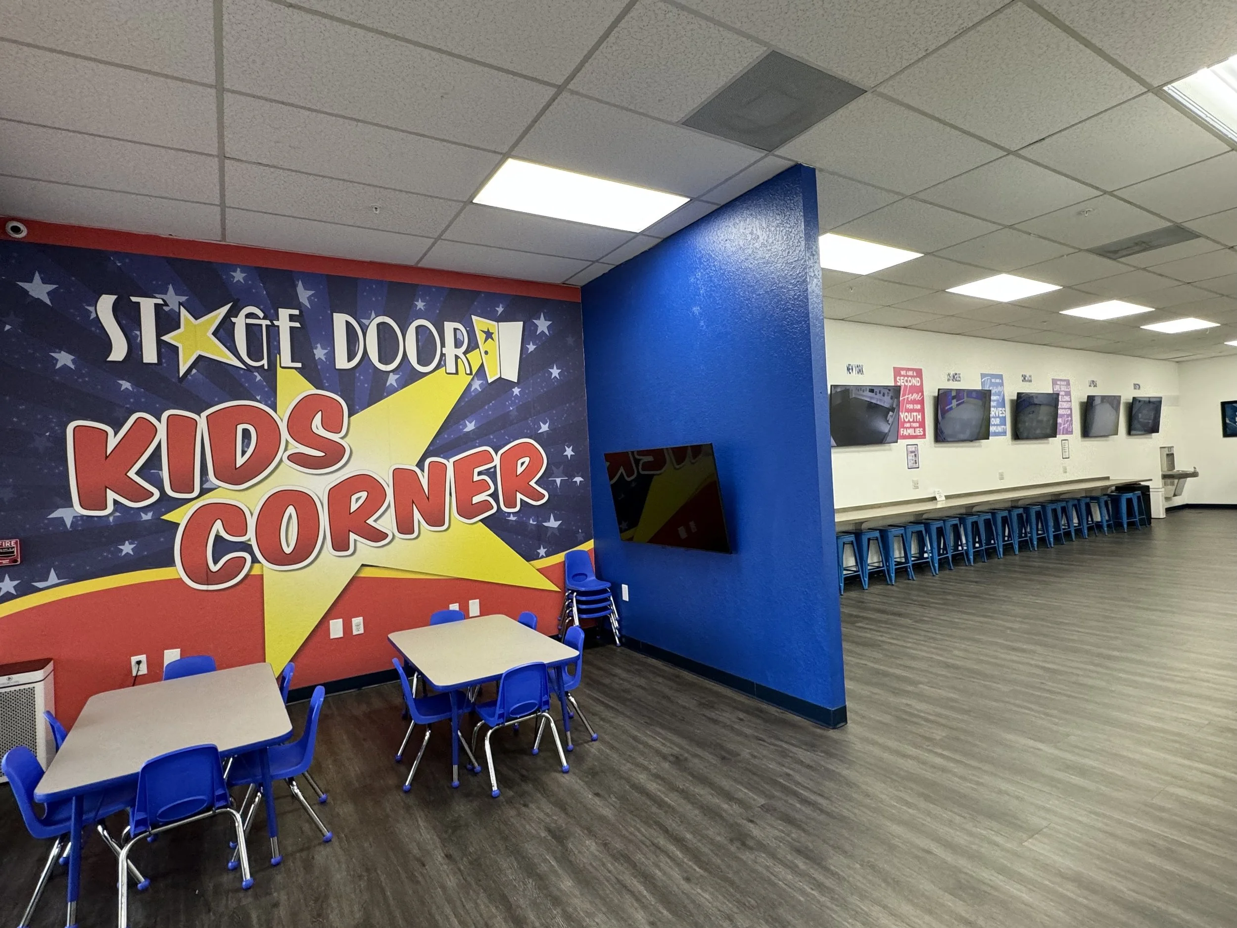 An indoor kids' corner with blue chairs and small tables near a colorful wall with stars and bold text.