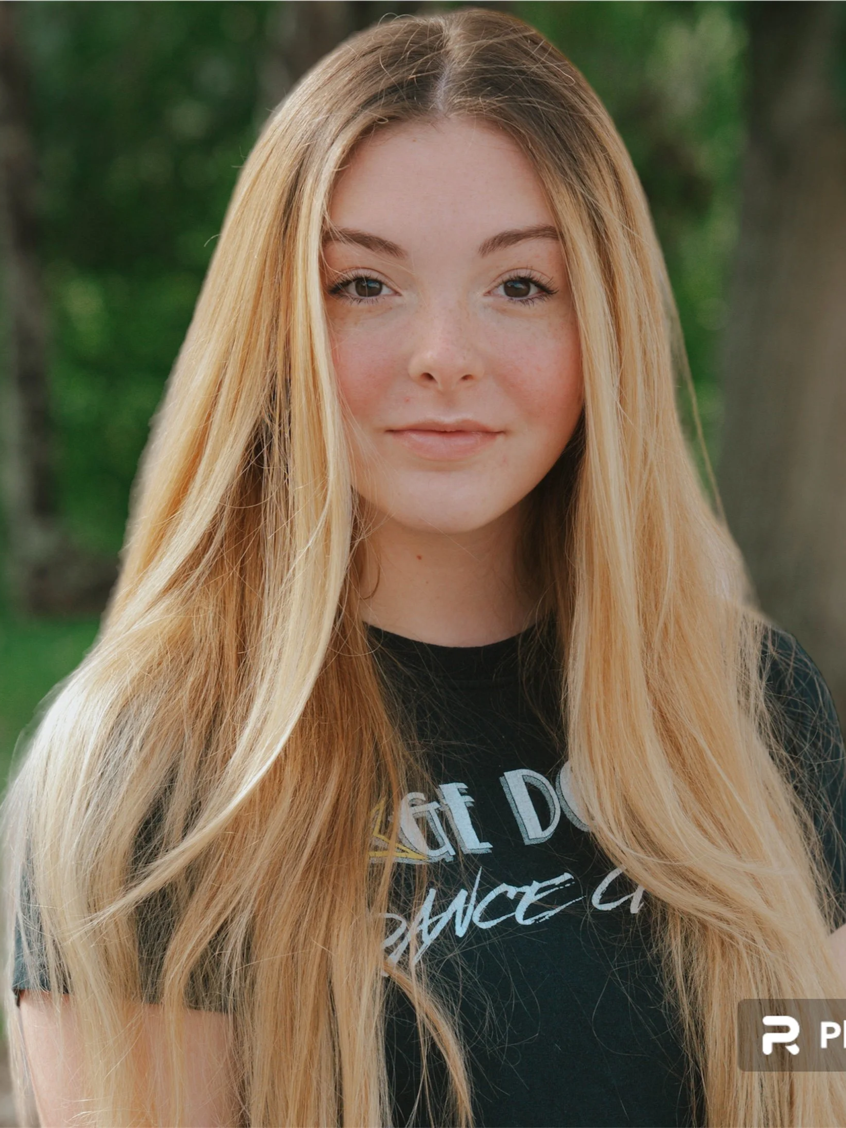 Close-up of a young woman with long, wavy blonde hair, wearing a black T-shirt with partially visible white text, standing outdoors with blurry green trees in the background.