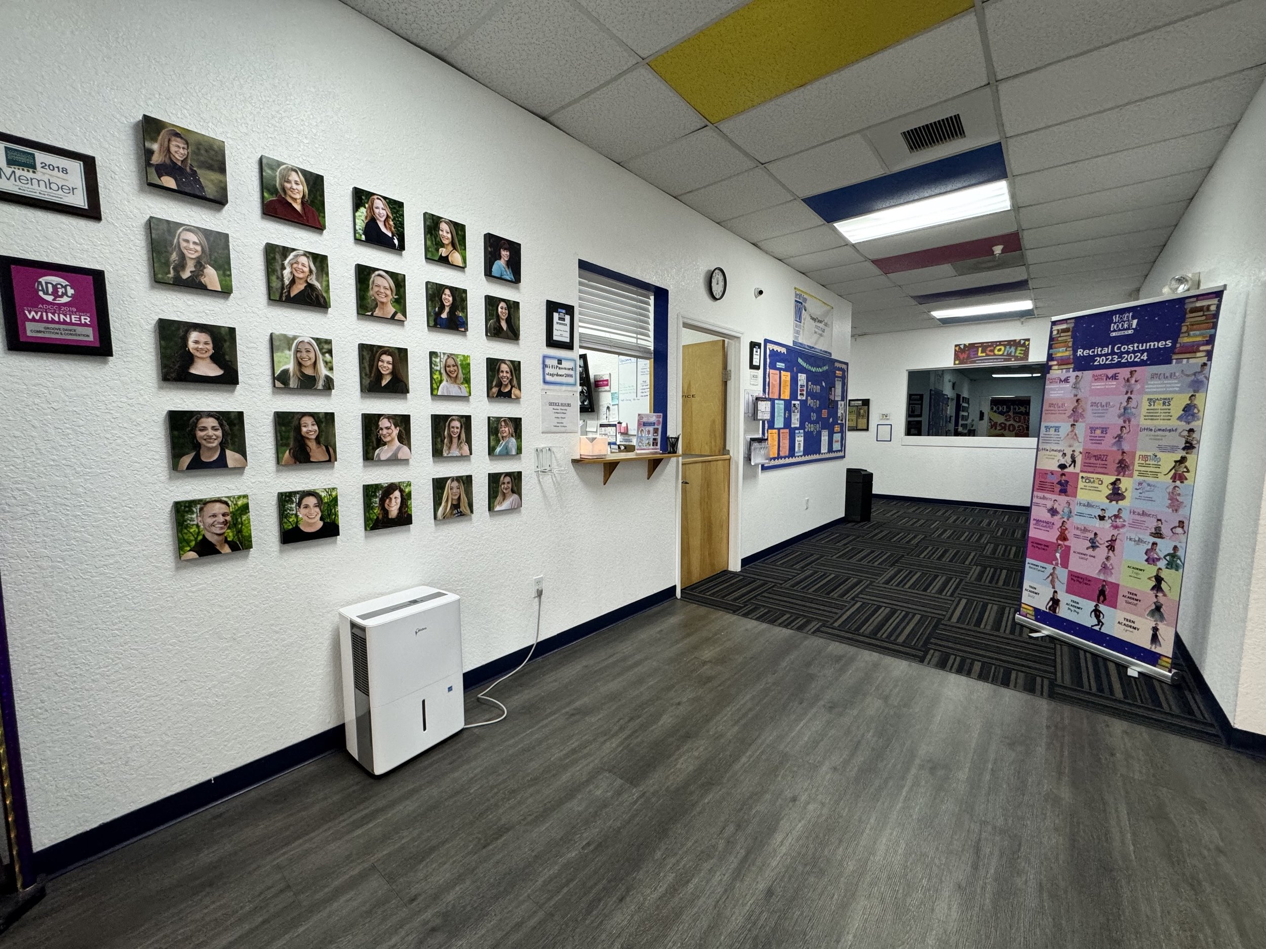 Indoor hallway with portraits of smiling young women on the wall, a sign-up sheet, and a exhibition display for recital costumes 2023-2024.