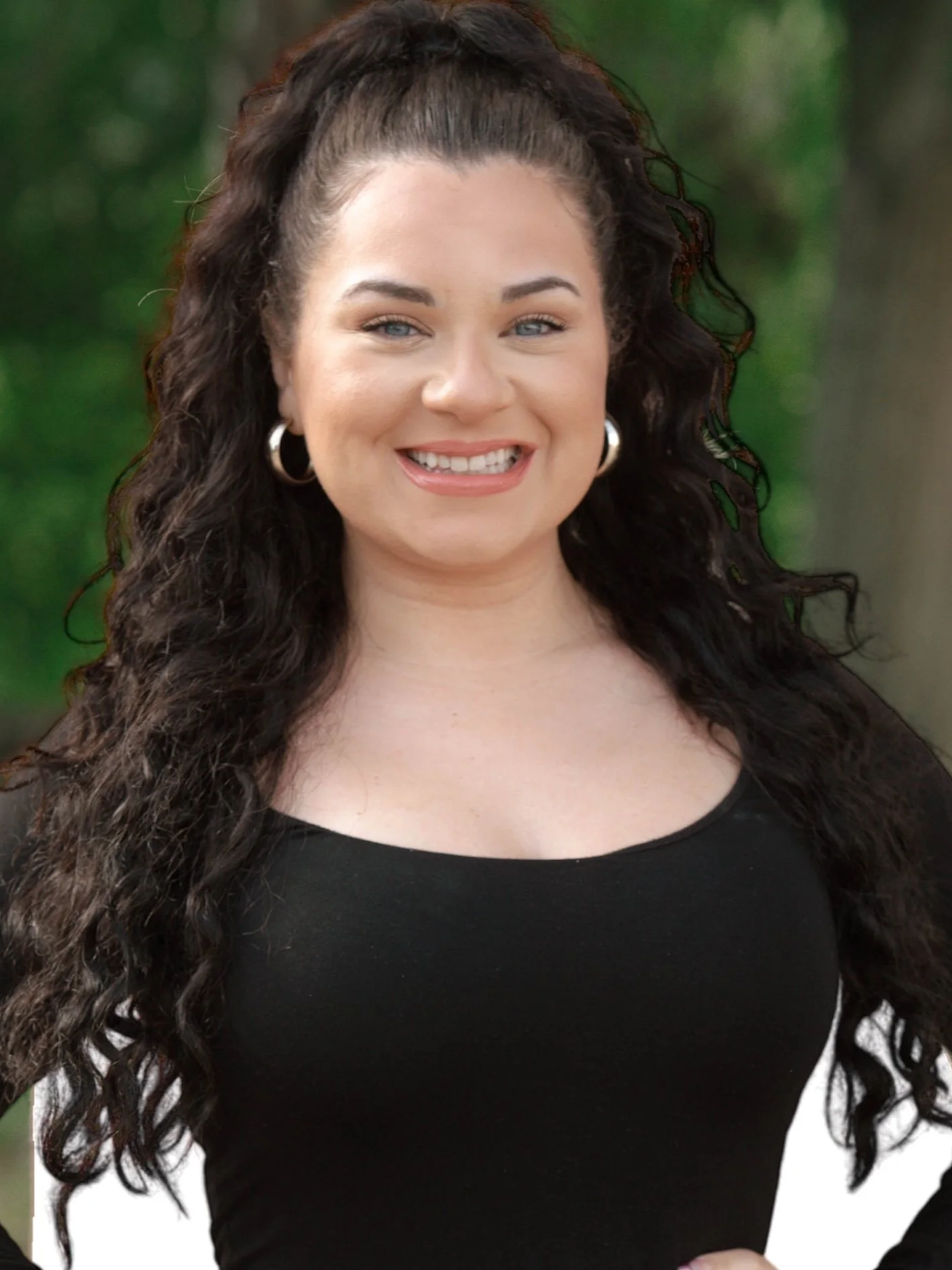 A woman with long curly brown hair tied up in a ponytail, wearing hoop earrings, a black top, smiling, with green foliage in the background.