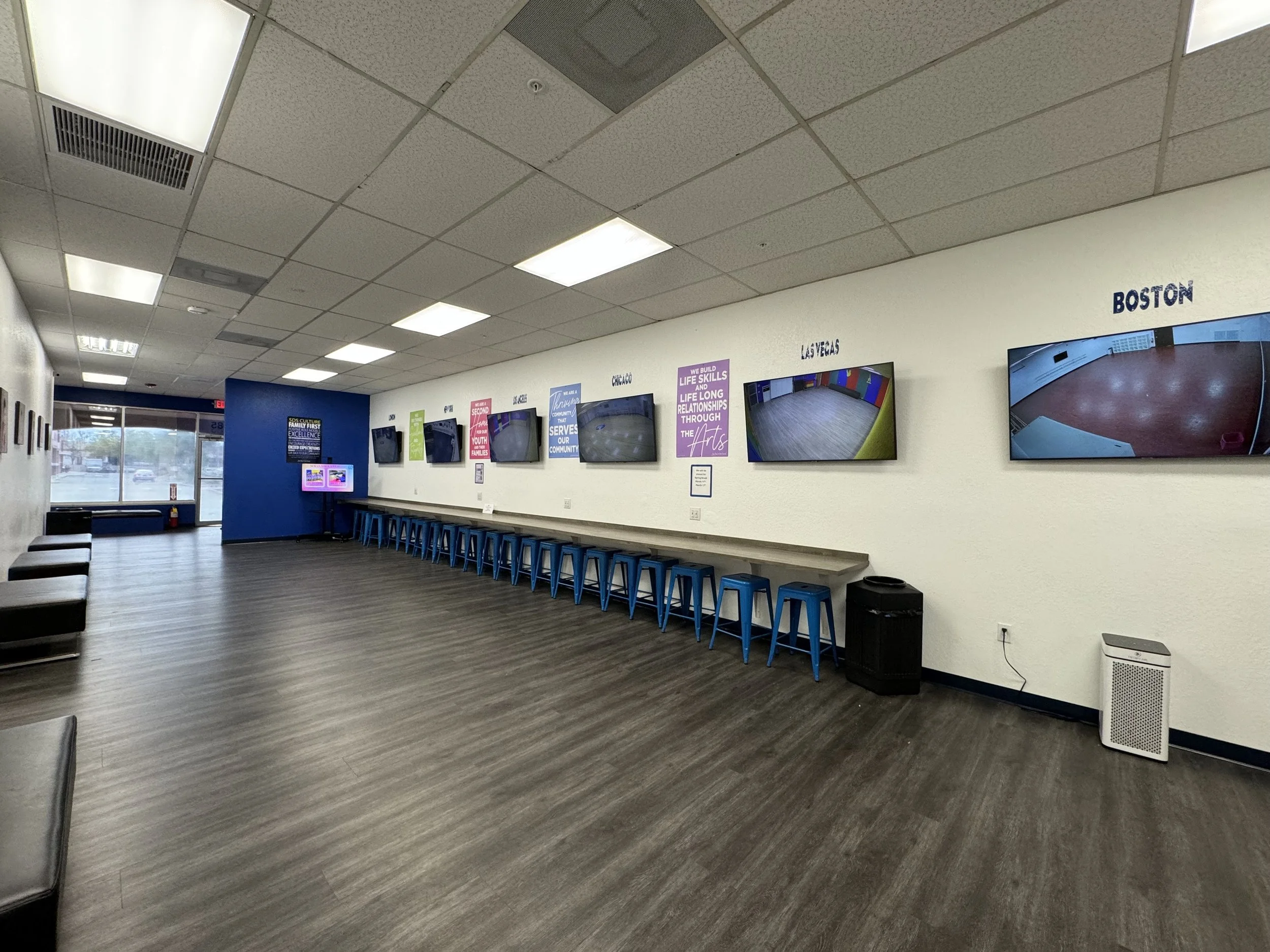 Empty waiting room with gray chairs, a row of blue stools, wall-mounted TVs, and posters on the wall, near a glass door and window.