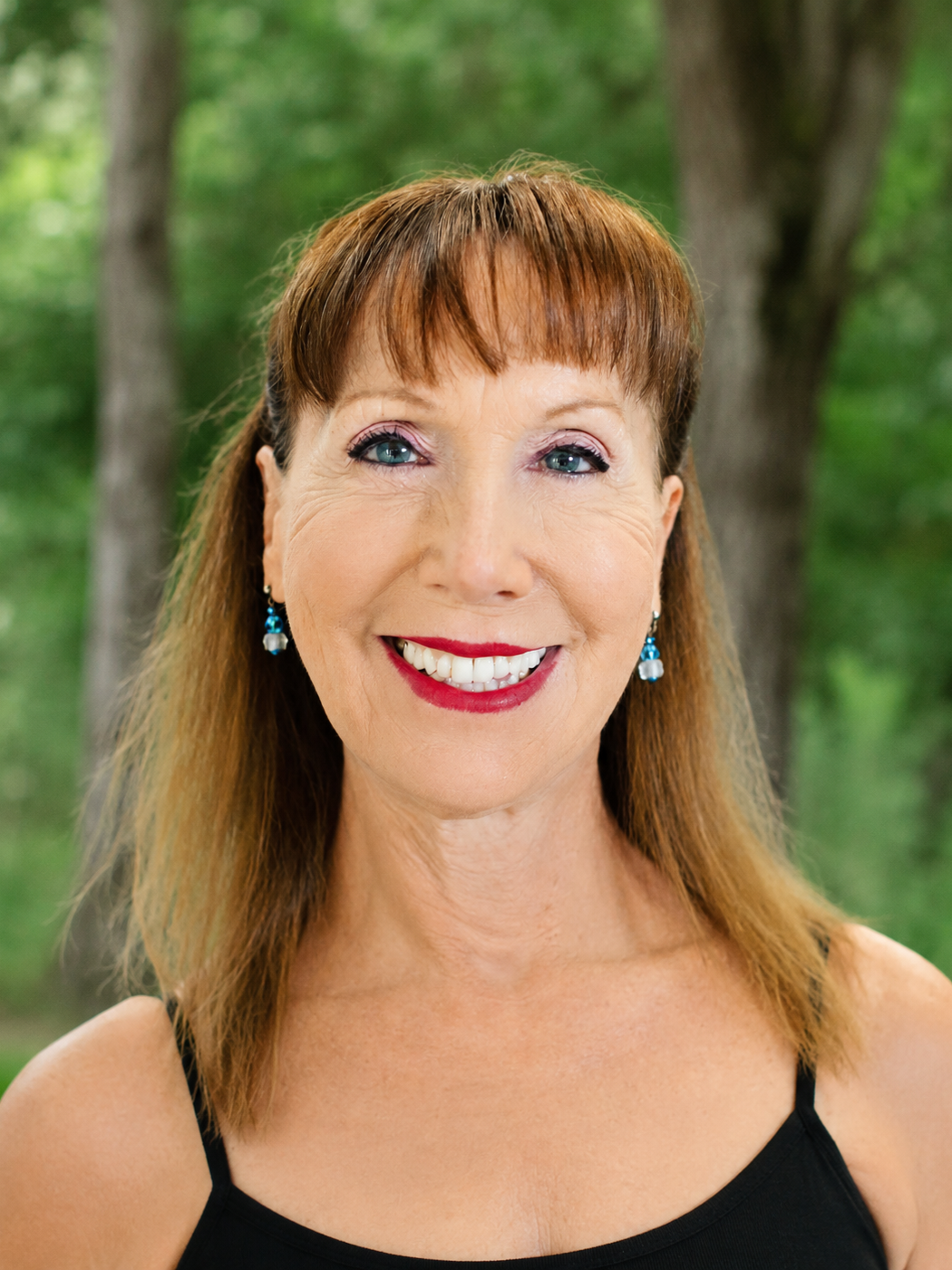 A smiling woman with red hair, wearing a black top and blue earrings, standing outdoors with trees and greenery in the background.