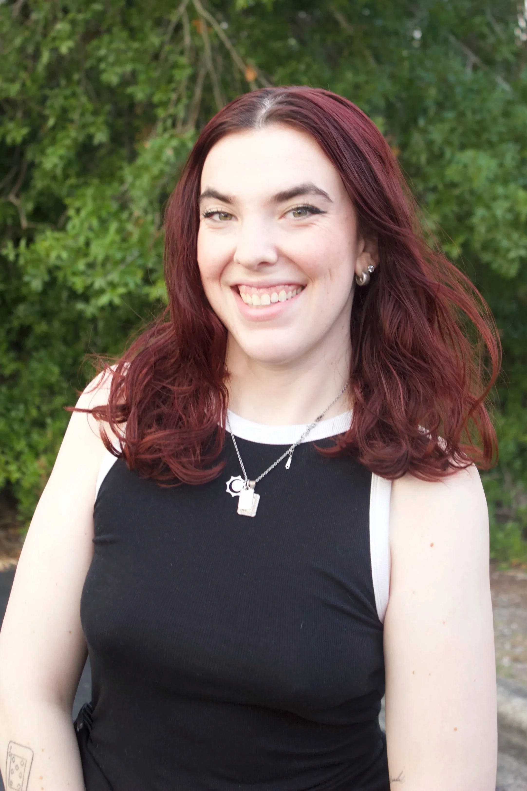 A young woman with long, curly red hair, wearing a black sleeveless top with white accents, silver earrings, and layered necklaces, smiling outdoors with green trees in the background.