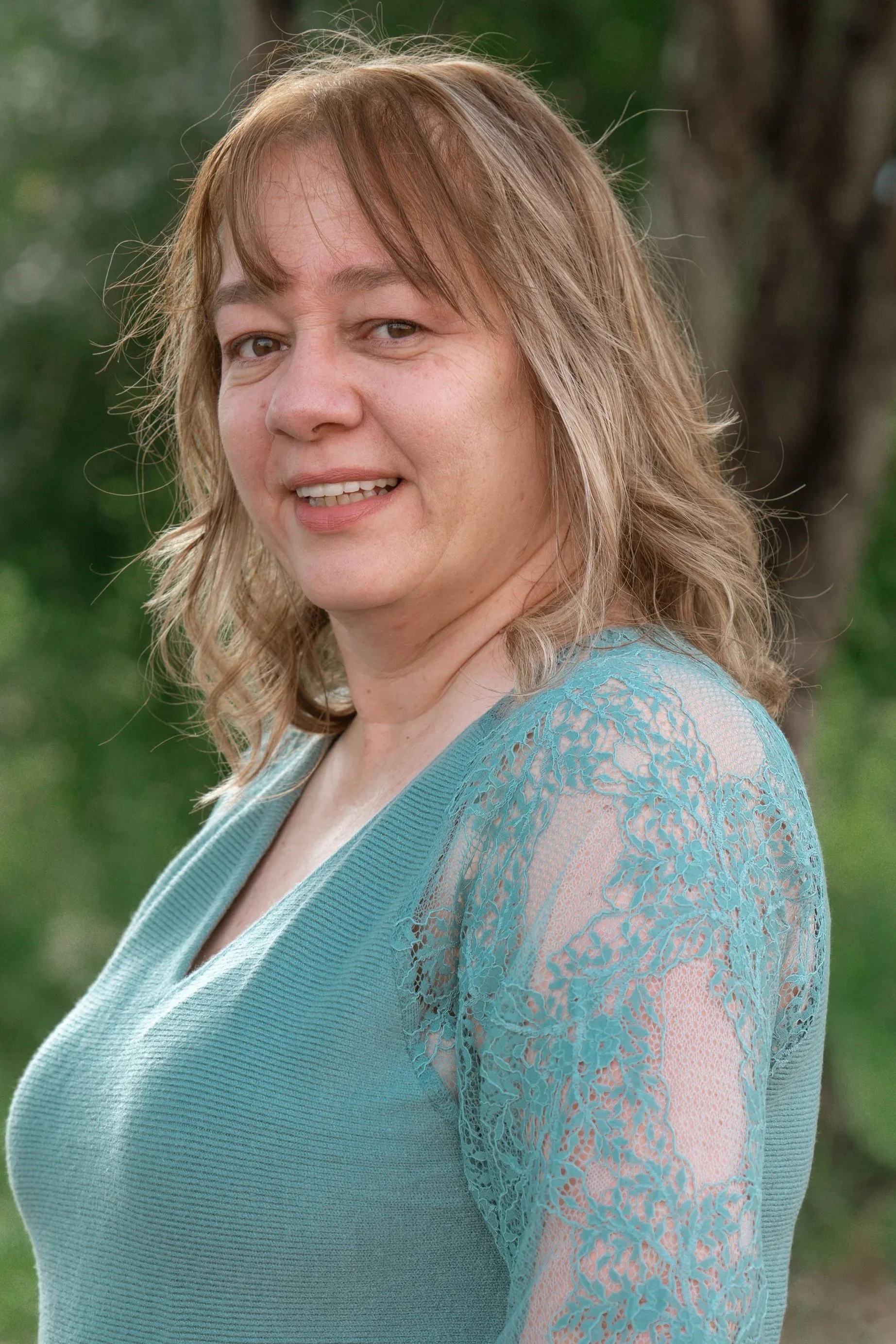 A woman with reddish-brown hair, smiling slightly, wearing a light blue lace dress, outdoors with green trees in the background.