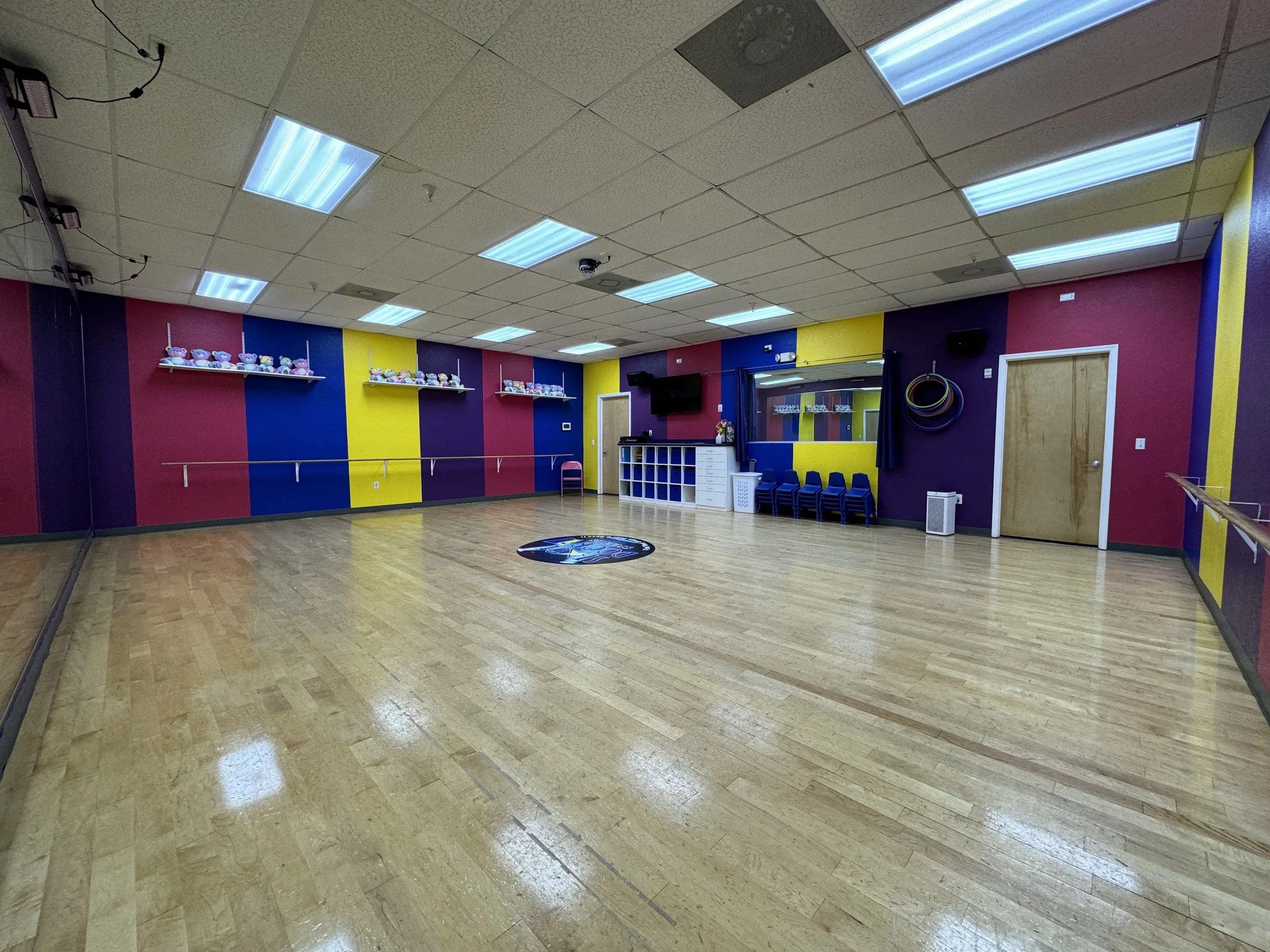 An indoor dance or exercise studio with colorful walls painted in pink, blue, and yellow. There is a mirrored wall on the right, a television mounted on the back wall, and shelves with plush toys on the left. The floor is wooden, and the ceiling has fluorescent lights.
