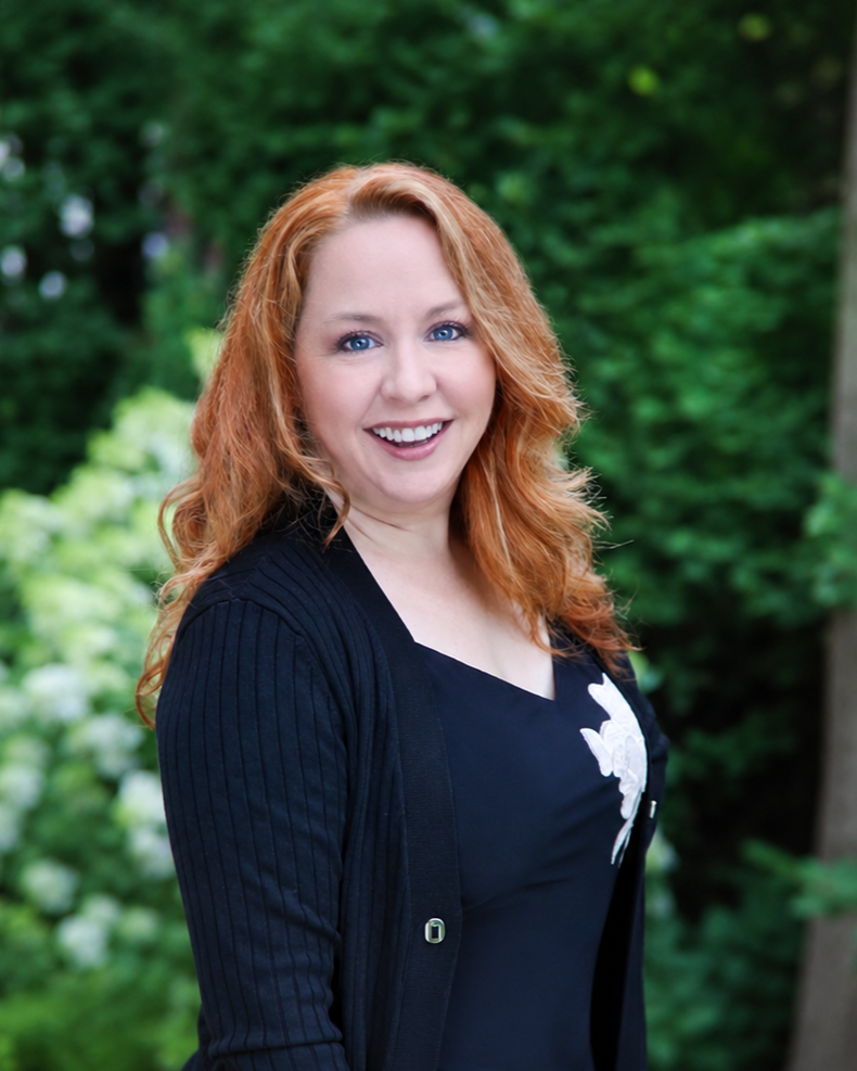 A smiling woman with curly red hair, wearing a black top and a black cardigan, standing outdoors in front of green trees.