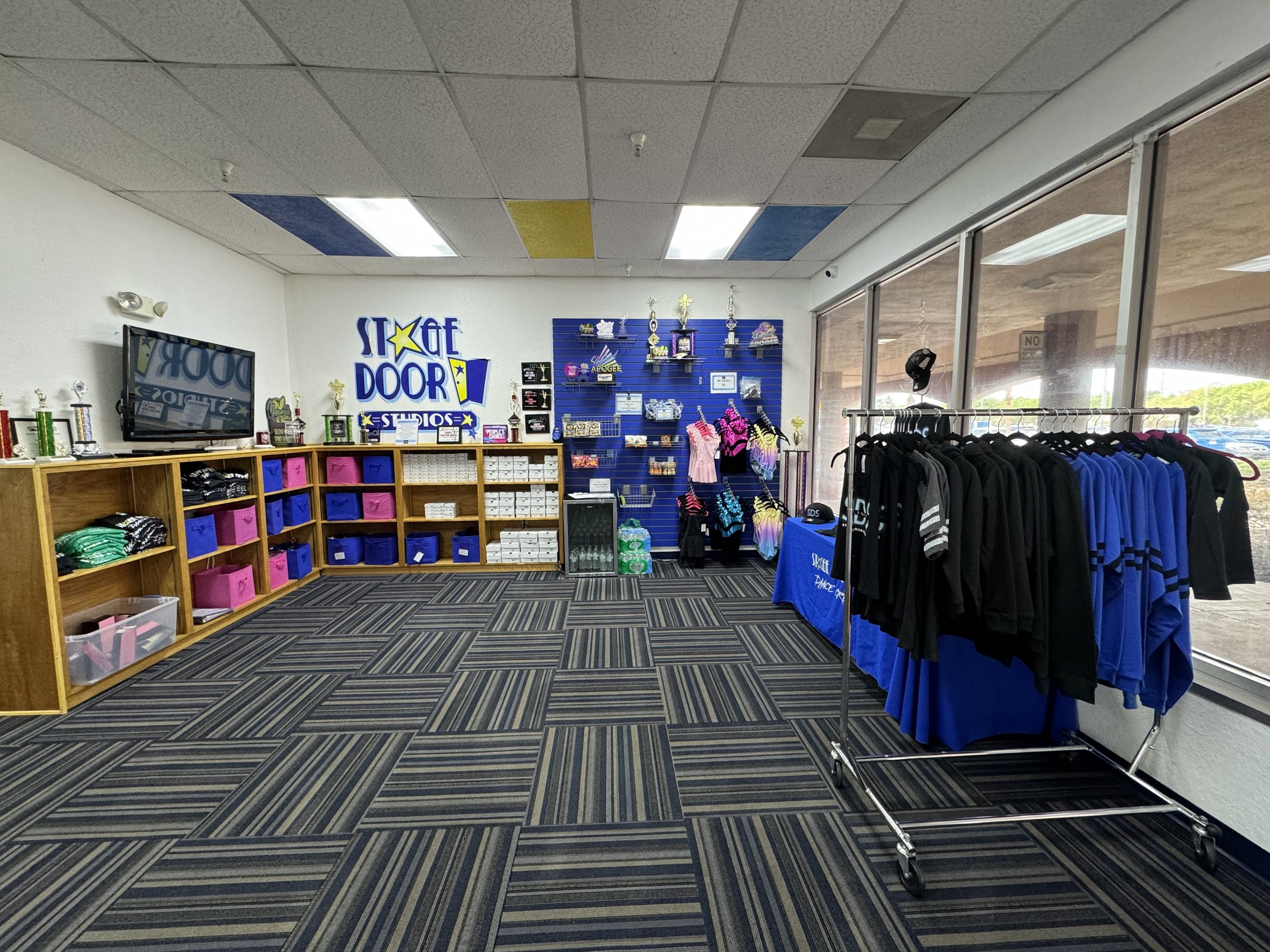 Interior of a retail store with blue and yellow ceiling tiles, a display rack of clothing, a wall with trophies and awards, and a large window showing the outside. Racks of apparel and a table with merchandise are visible.