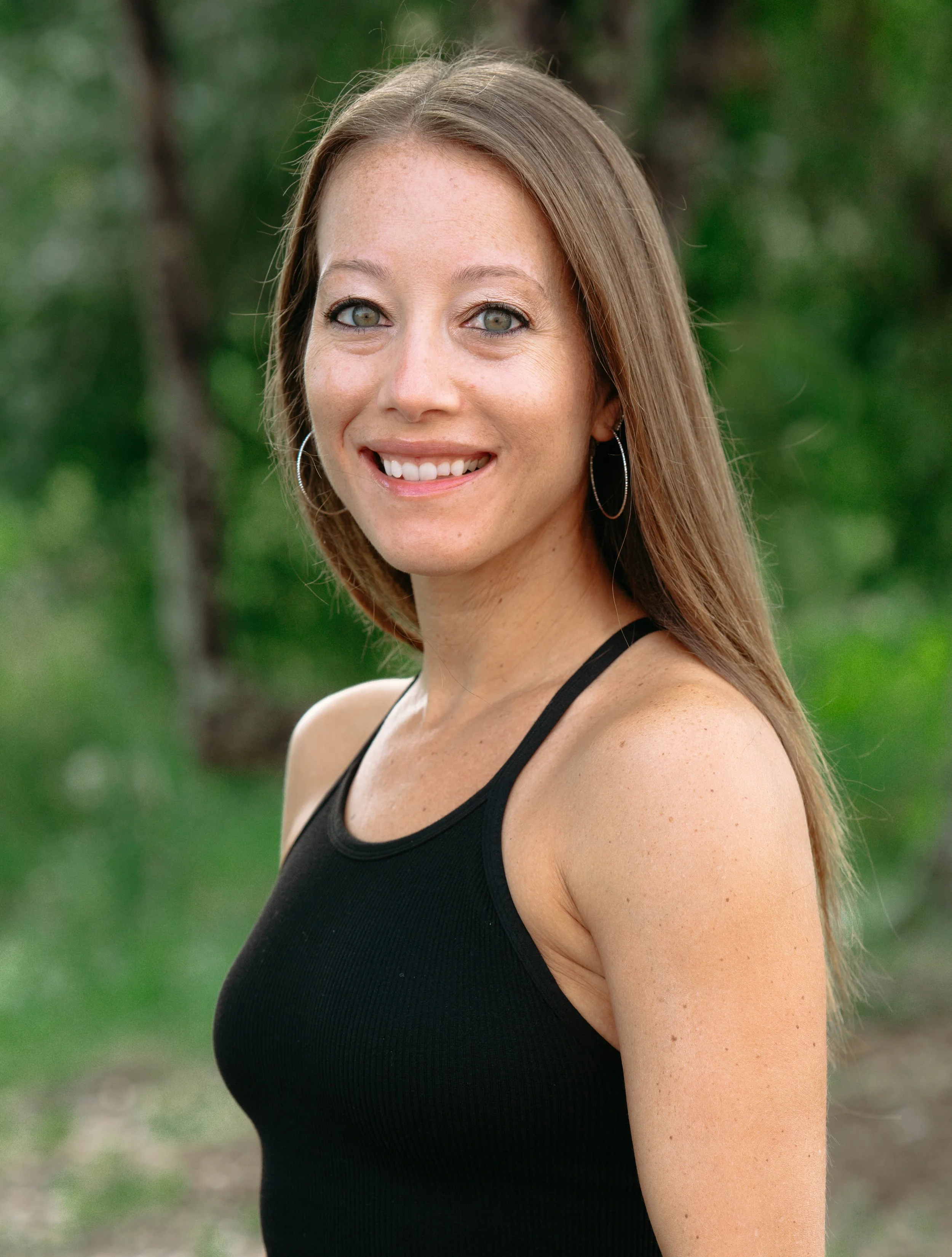 Close-up of a woman with long, light brown hair wearing a black tank top, standing outdoors with a blurred green natural background, smiling and looking at the camera.