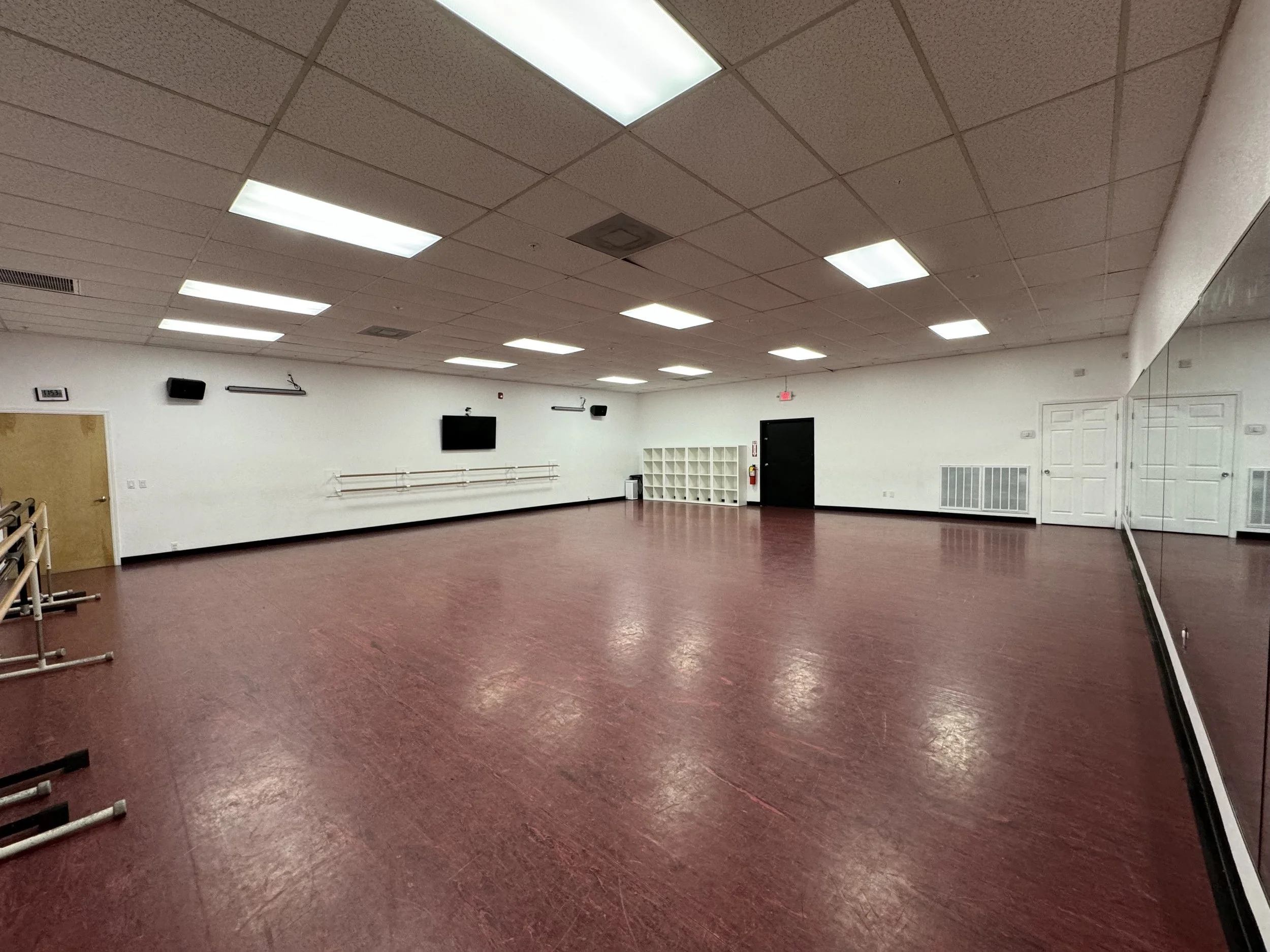 An empty dance studio with a maroon floor, white walls, fluorescent ceiling lights, a large mirror on one side, wall-mounted speakers, a wall-mounted television, ballet barres along the wall, a wall clock, a door with an exit sign, and a white cubby 
