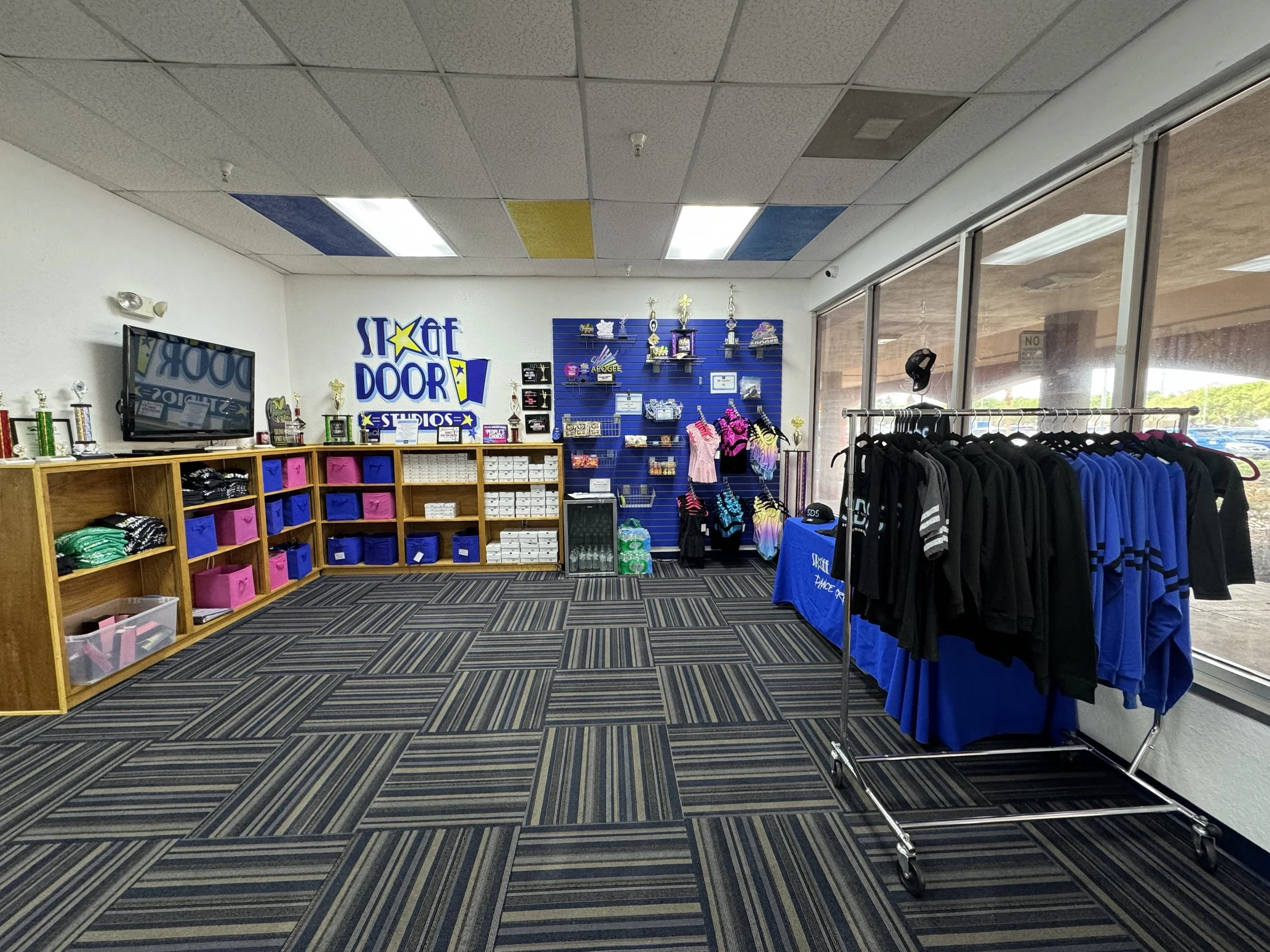 Interior of a sports apparel store with clothing racks, a display table, wall shelves with trophies, and blue, yellow, and white decor.