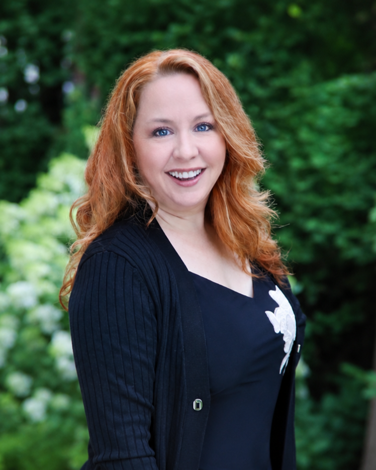 A smiling woman with red hair and blue eyes standing outdoors in front of green foliage.