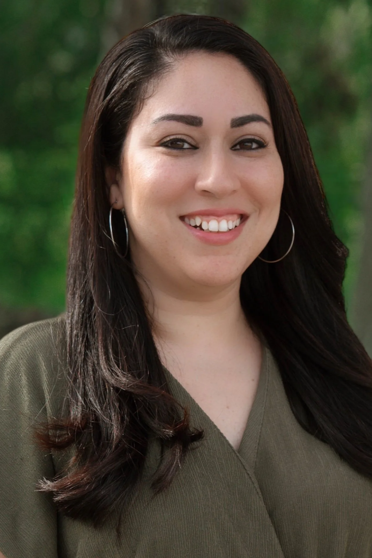 A woman smiling outdoors with long, dark hair and hoop earrings, wearing a green top, with green trees in the background.