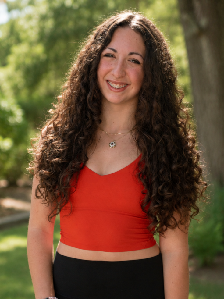 Young woman with long curly hair, smiling, wearing a red crop top and black skirt, standing outdoors in a park with green trees.