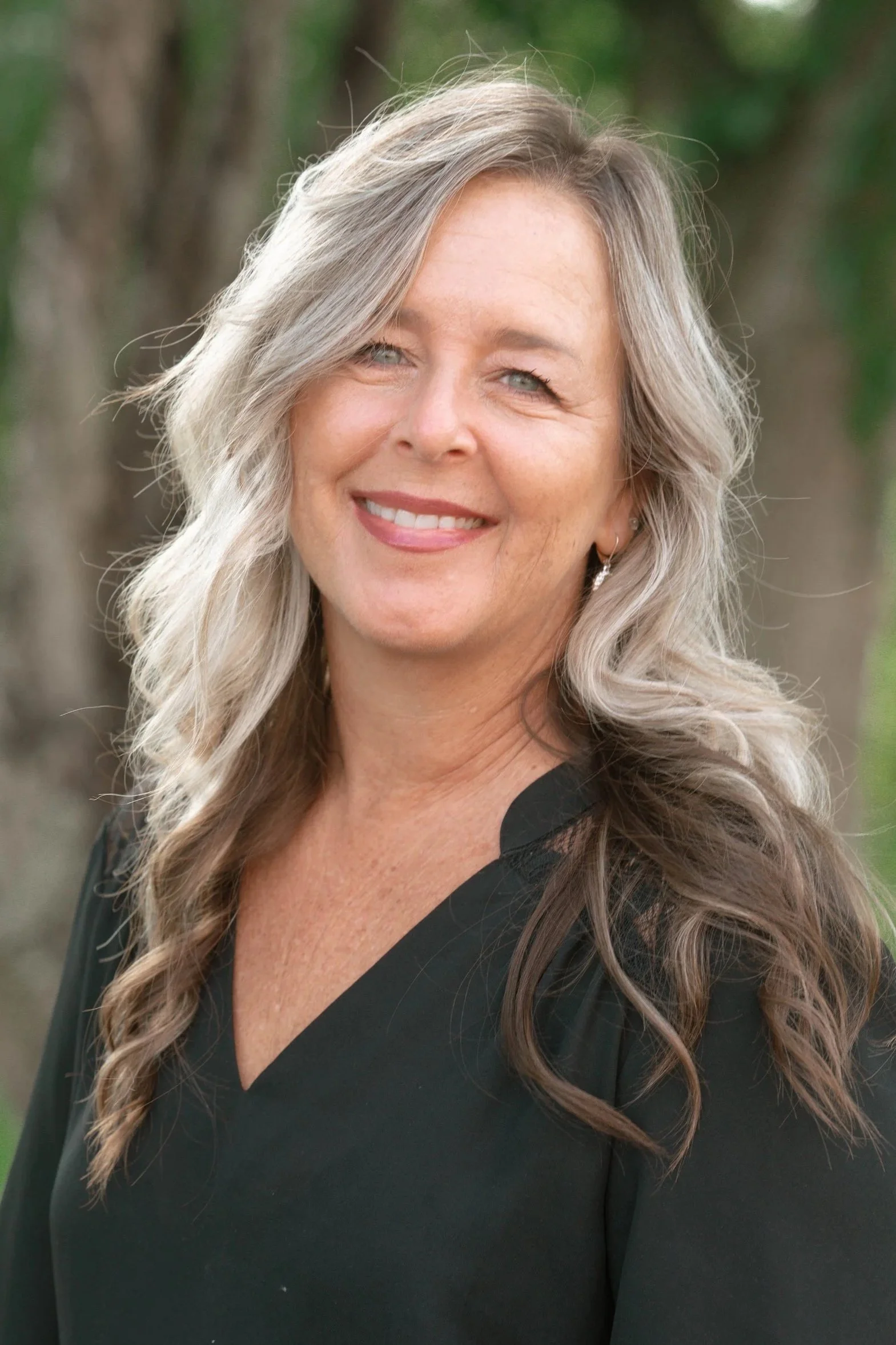 A woman with long, wavy blonde hair and blue eyes, smiling outdoors with a blurred green background, wearing a black top.