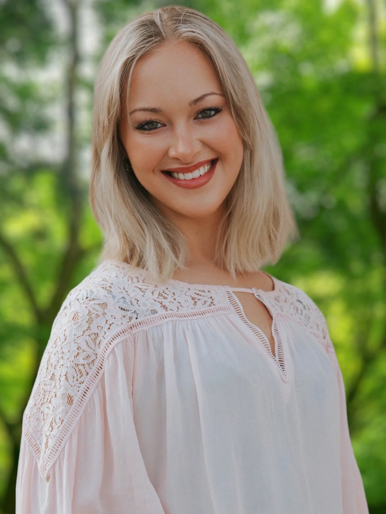 A woman with blonde hair, green eyes, and a bright smile wearing a white dress with lace detailing, standing outdoors with green trees in the background.