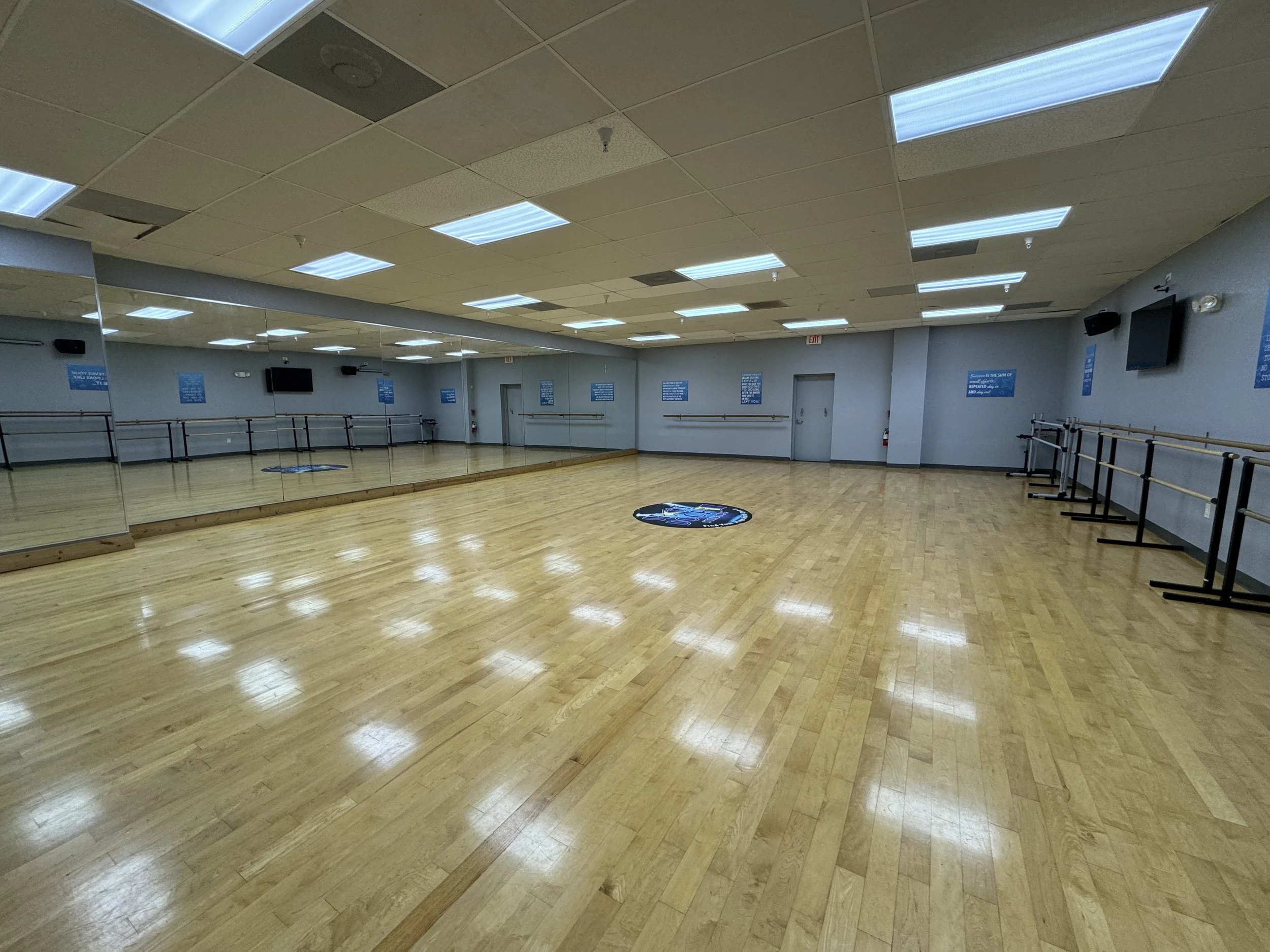 An empty dance studio with wooden floors, large mirrors along one wall, and ceiling lights. There are blue signs on the walls, a mounted TV, and ballet barres along the mirrored wall.