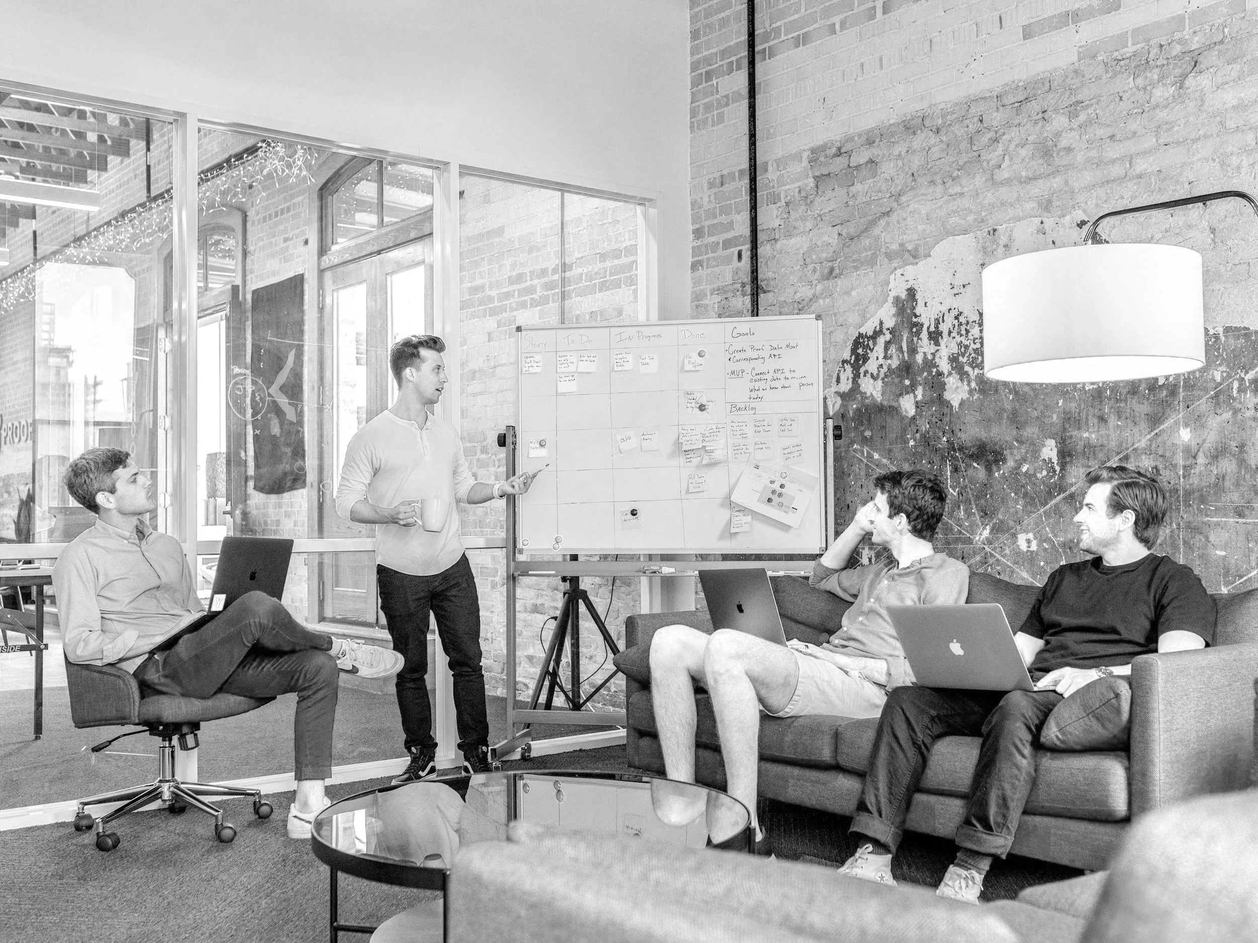 A group of young designers in a modern office space with exposed brick walls, some seated on chairs and a sofa, while one stands at a whiteboard giving a presentation. There are laptops, a laptop on a chair, and a coffee table in the foreground.